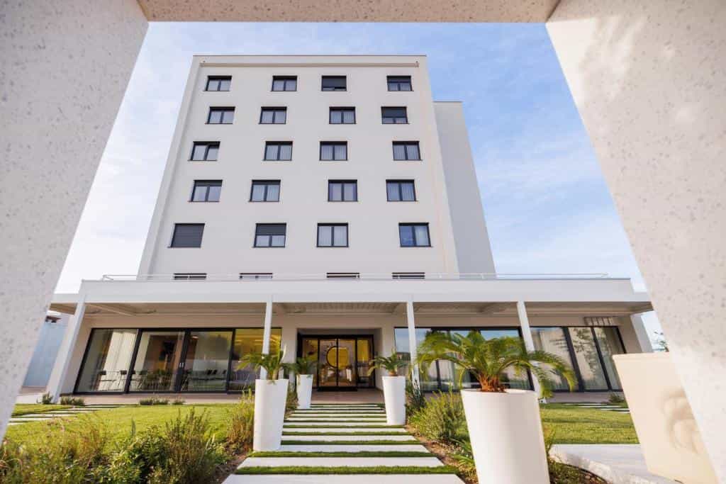 Modern white hotel building entrance framed by white architectural elements and green landscaping.