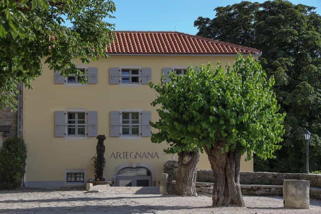 Yellow building with "ARTEGNANA" sign, trees, and cobblestone courtyard