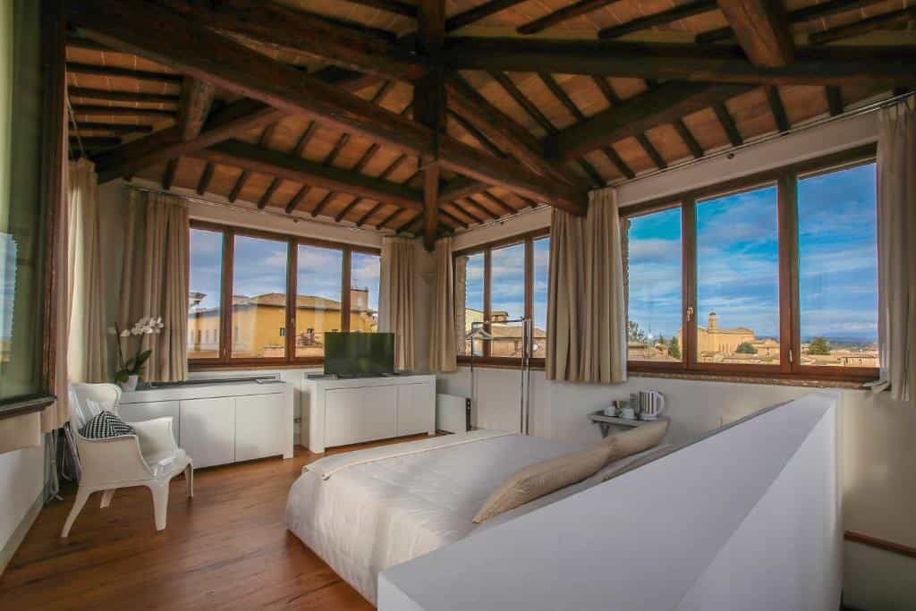 Attic bedroom with exposed wood beams, white furniture, and town view through many windows.