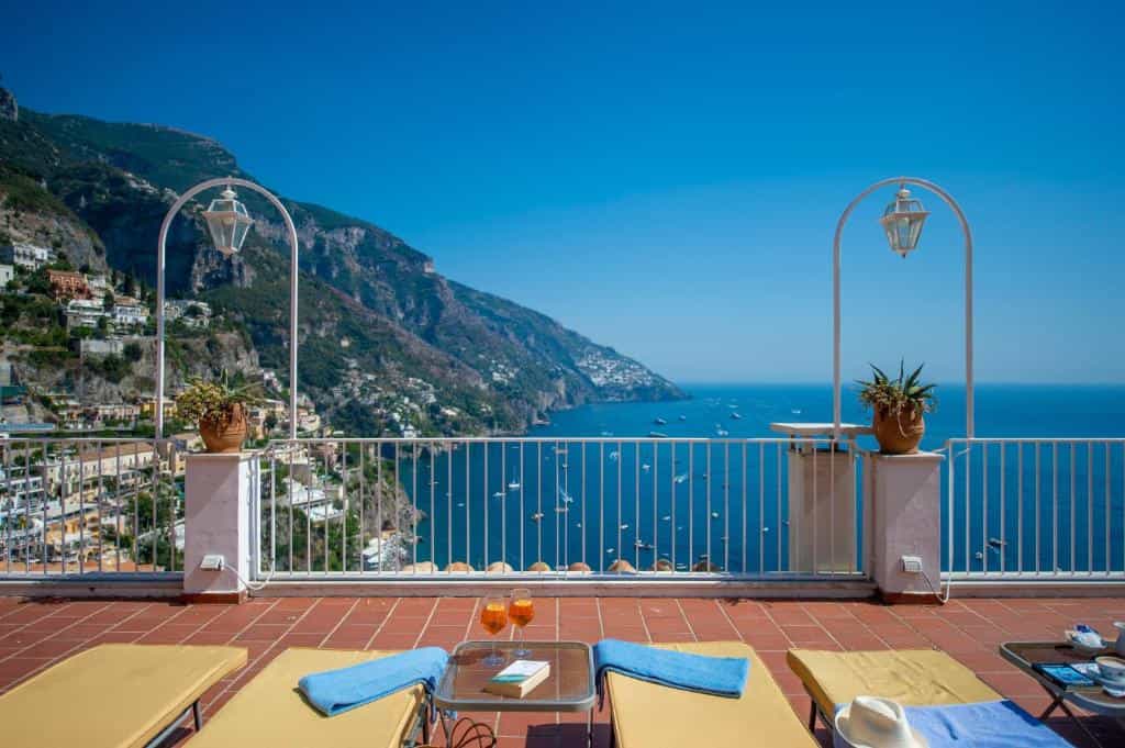 Terrace with sun loungers, drinks, and view of steep coastline and boats in the sea.