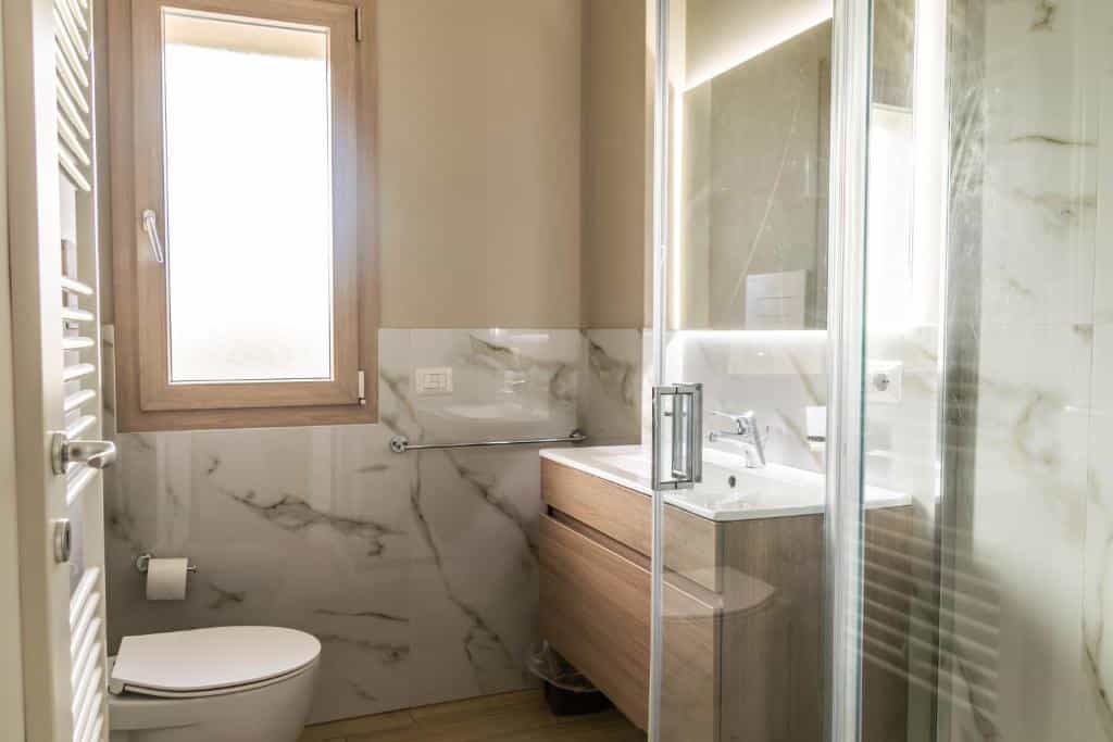 Modern bathroom with white marble tile, wood vanity, toilet, and glass shower enclosure.
