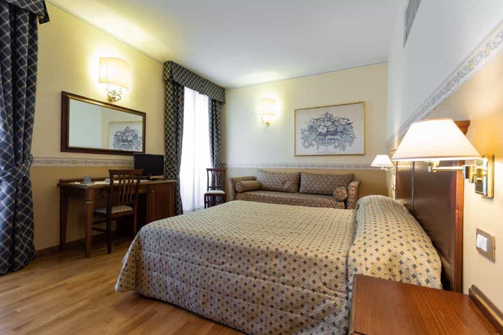 Hotel room with large bed, patterned sofa, wooden desk, and wall art.