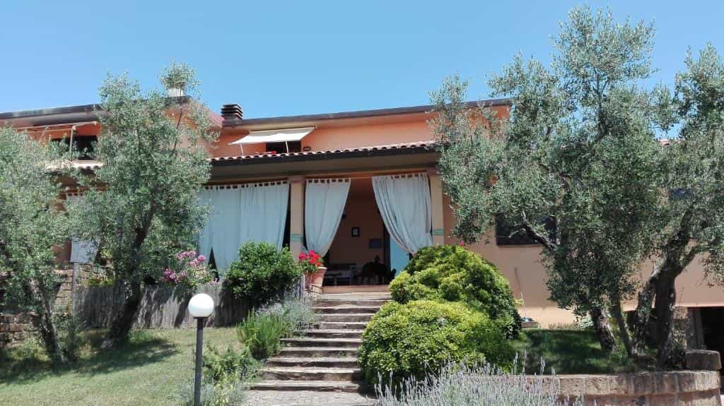 Peach-colored house entrance with stone steps, olive trees, and green bushes under a clear blue sky.