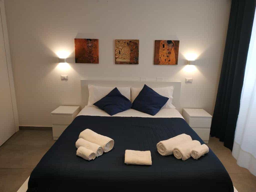 Double bed with navy quilt, white linens, and rolled towels in a room with Klimt art prints.
