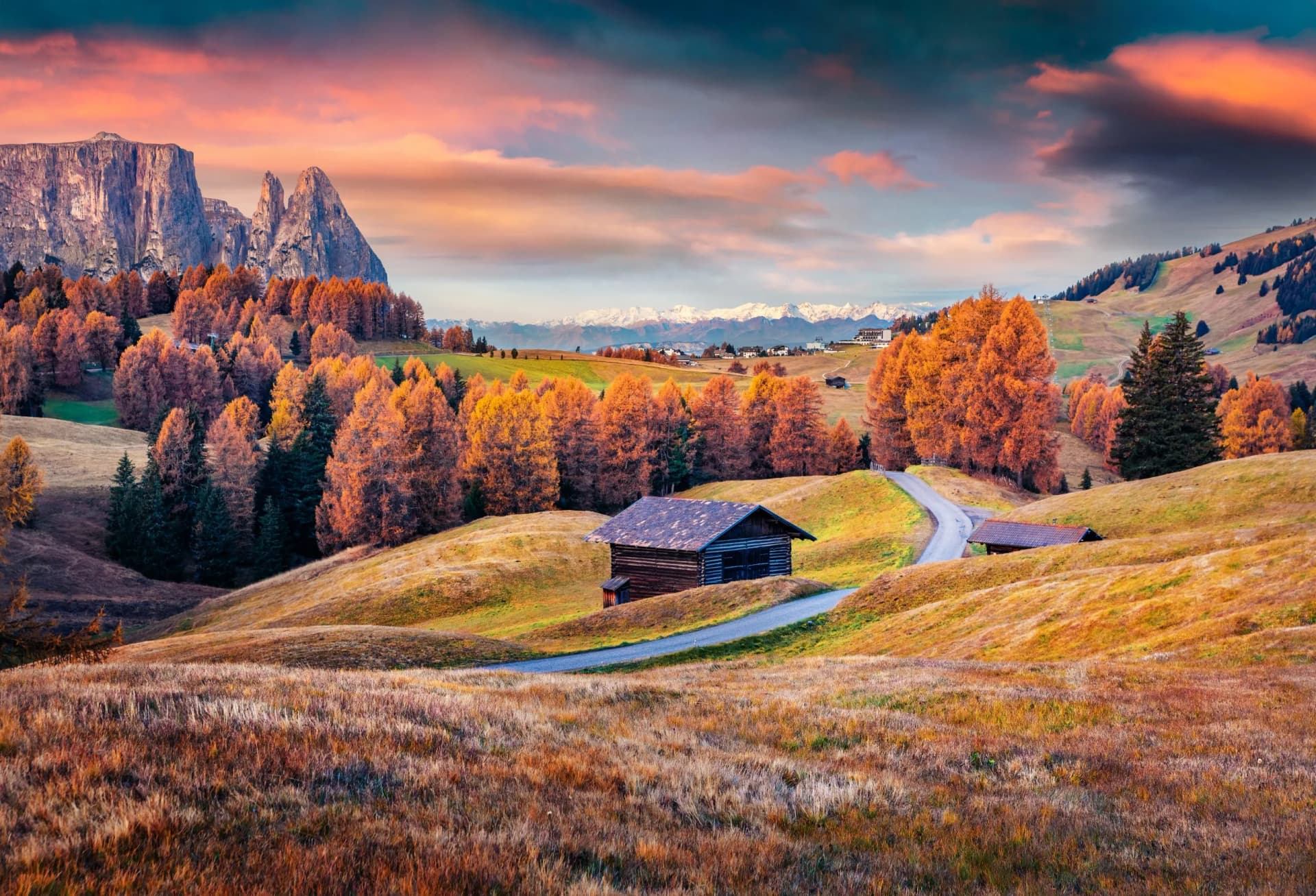 Seiser Alm mountain pastures with wooden huts, autumn trees, and snow-capped mountains at sunrise.