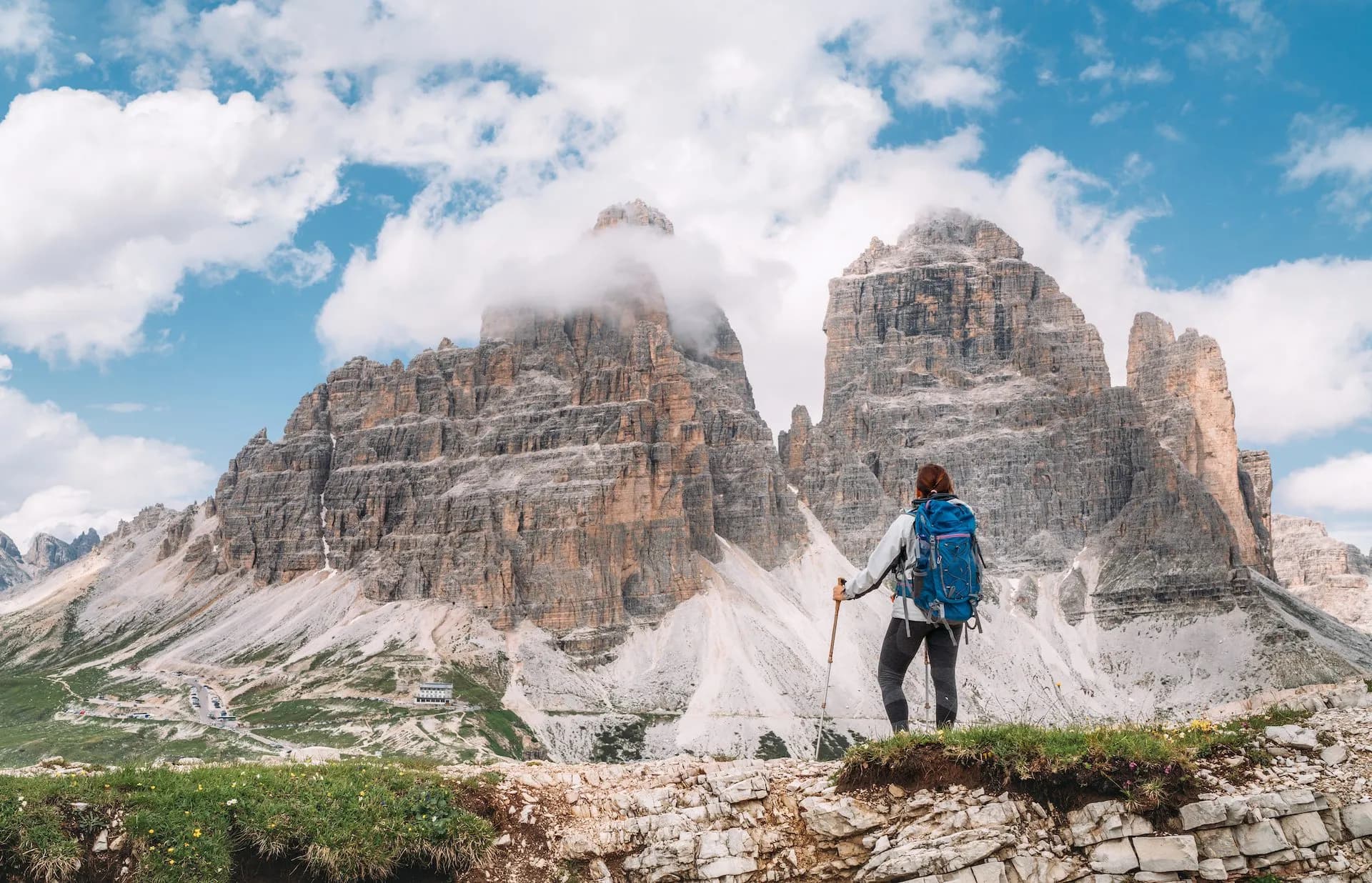Hiker with backpack and poles overlooks Tre Cime di Lavaredo peaks under cloudy sky.