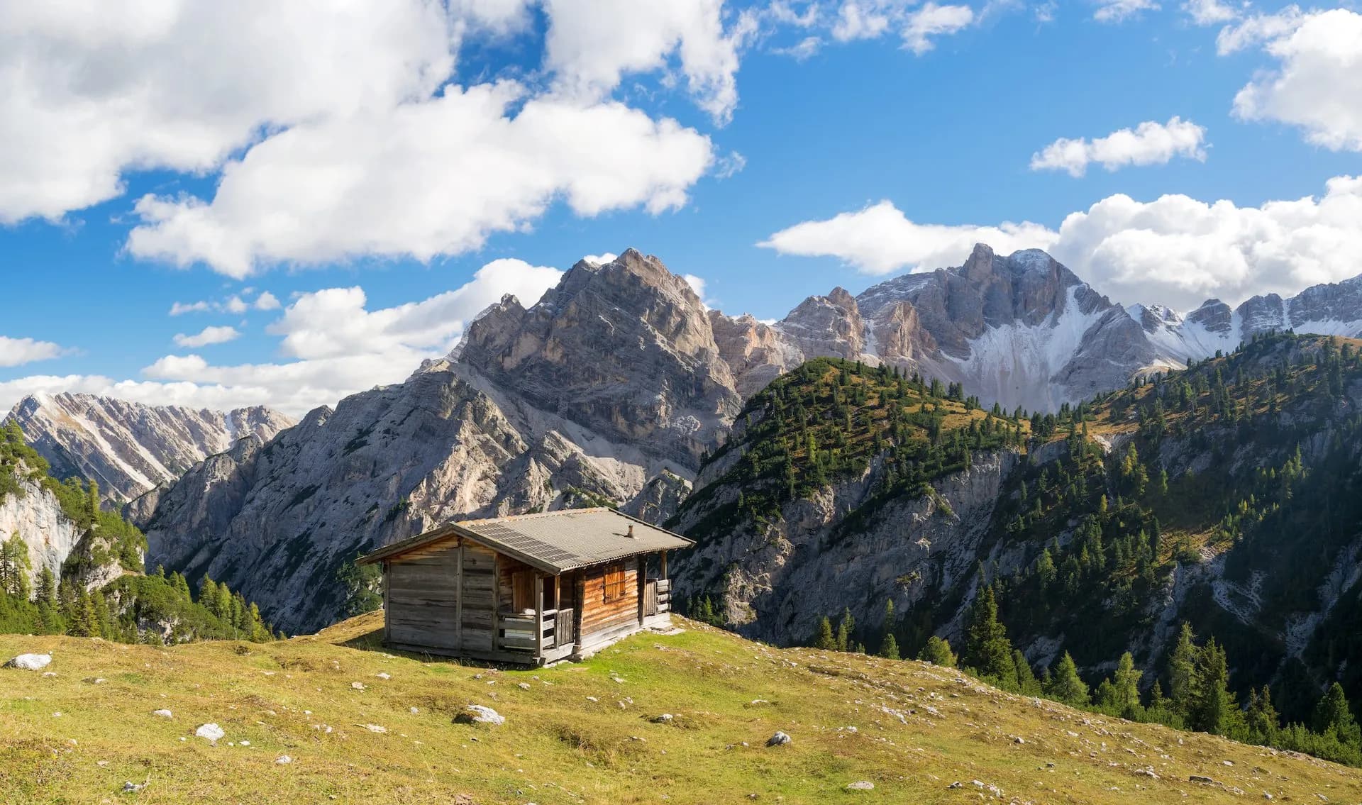 Wooden alpine hut on grassy slope with rugged, snow-dusted mountains under blue sky.