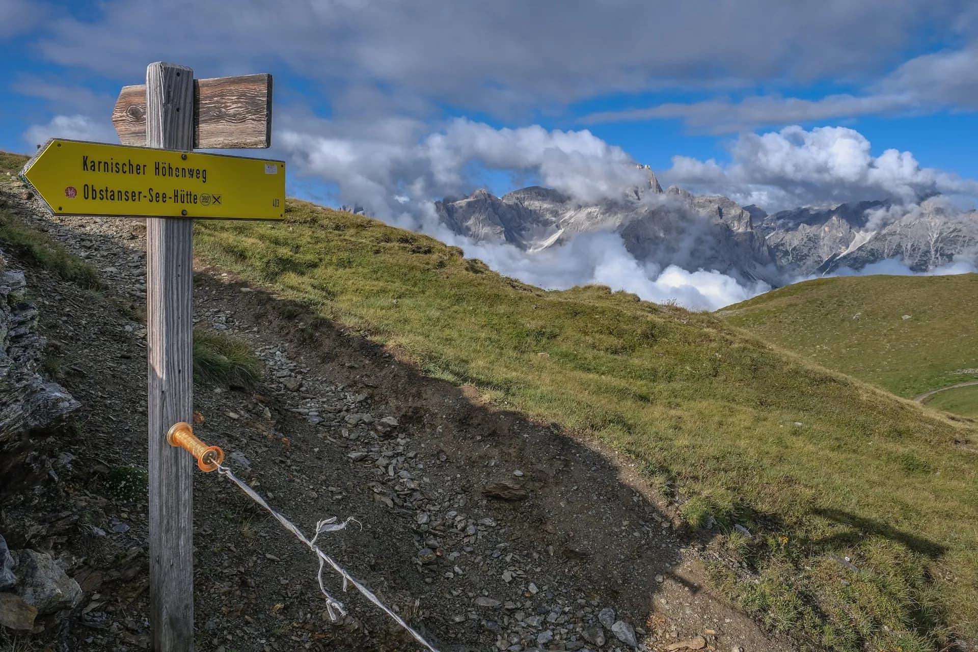 Hiking signpost on grassy slope pointing toward Karnischer Höhenweg and Obstanser-See-Hütte with mountains.