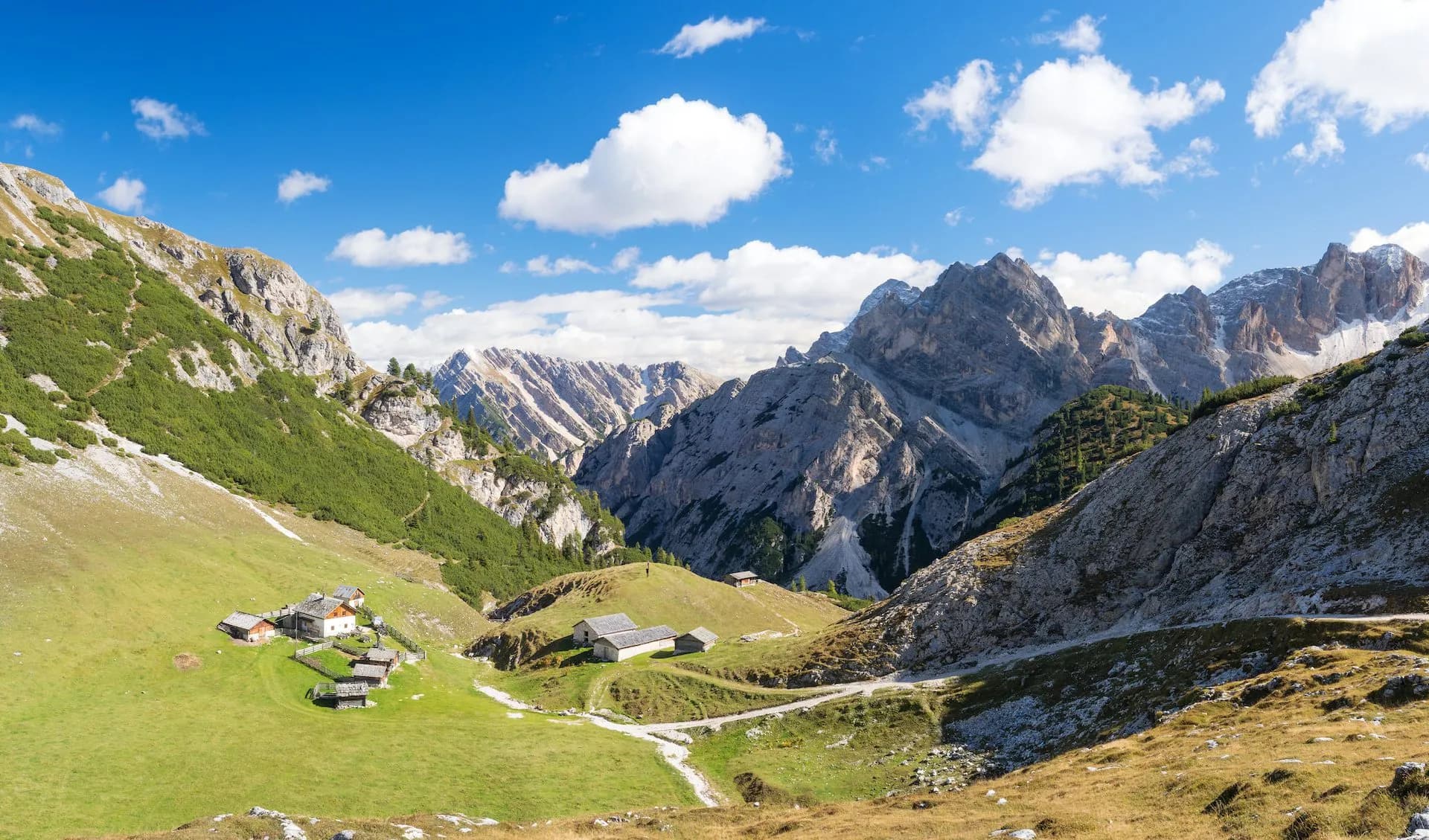 Alpine valley with green meadows, small farm buildings, and rugged mountains under a blue sky.