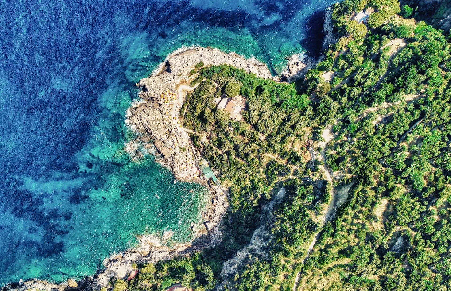 Aerial view of rocky coastline, lush green trees, and clear turquoise Mediterranean waters at Punta Campanella.