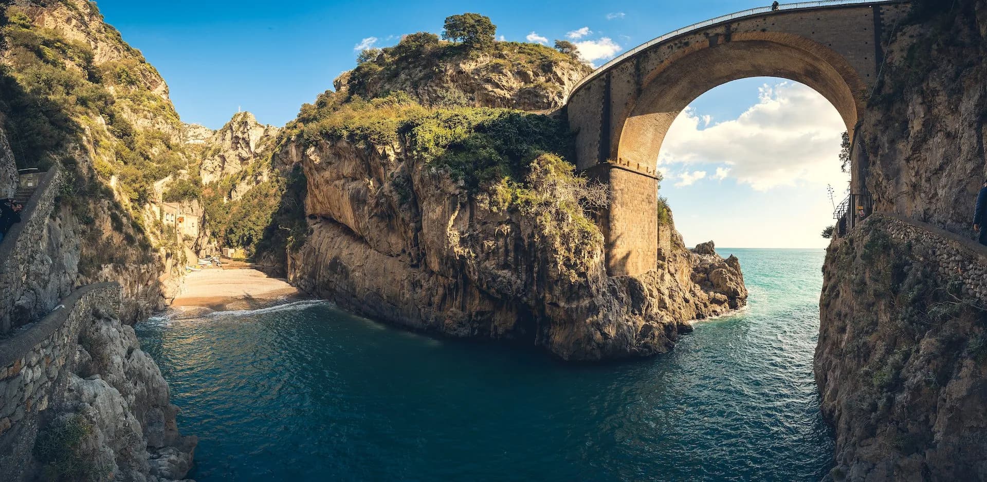 Stone arch bridge over turquoise sea inlet leading to a small beach between steep cliffs.