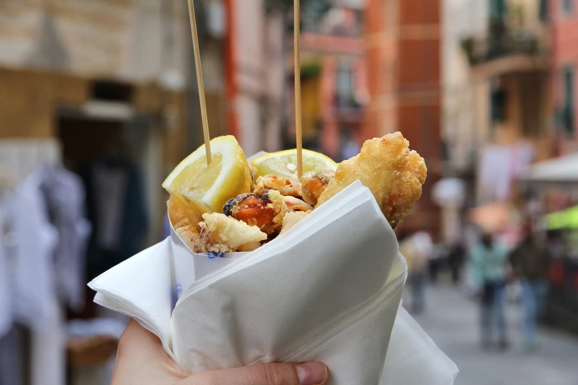 Hand holding paper cone of traditional fried seafood with lemon wedges on a city street.