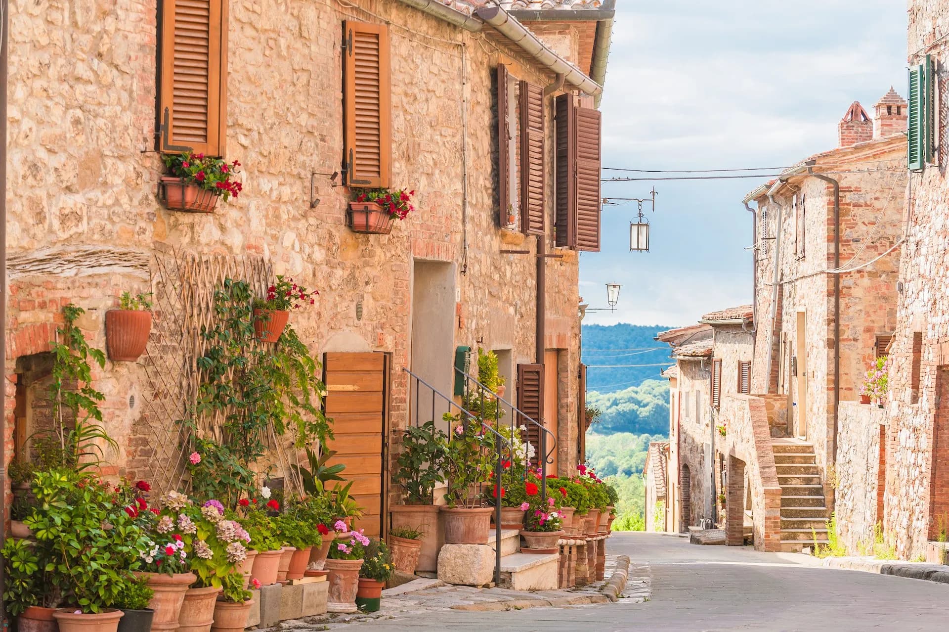 Stone street in Siena with flower pots lining rustic buildings and green hills in background.