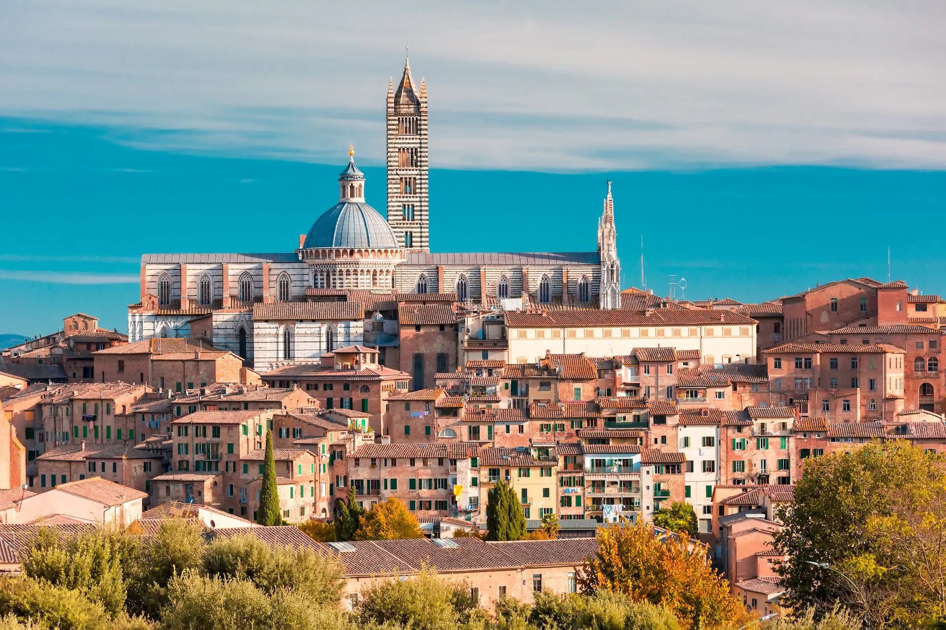 Siena cityscape with Duomo dome and striped bell tower over terracotta roofs under blue sky.
