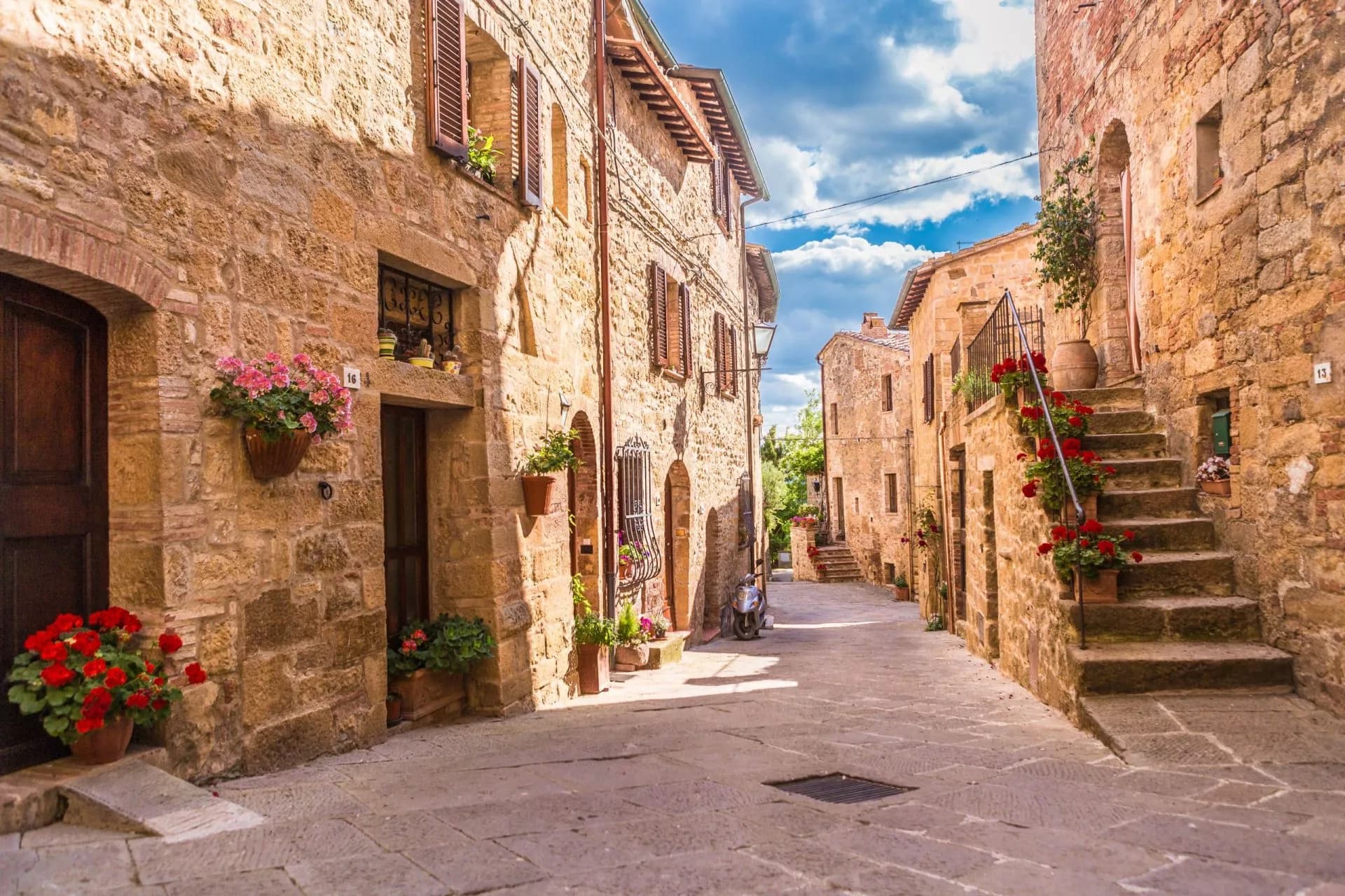 Cobblestone street in a historic Tuscan town with stone buildings and potted red and pink flowers.