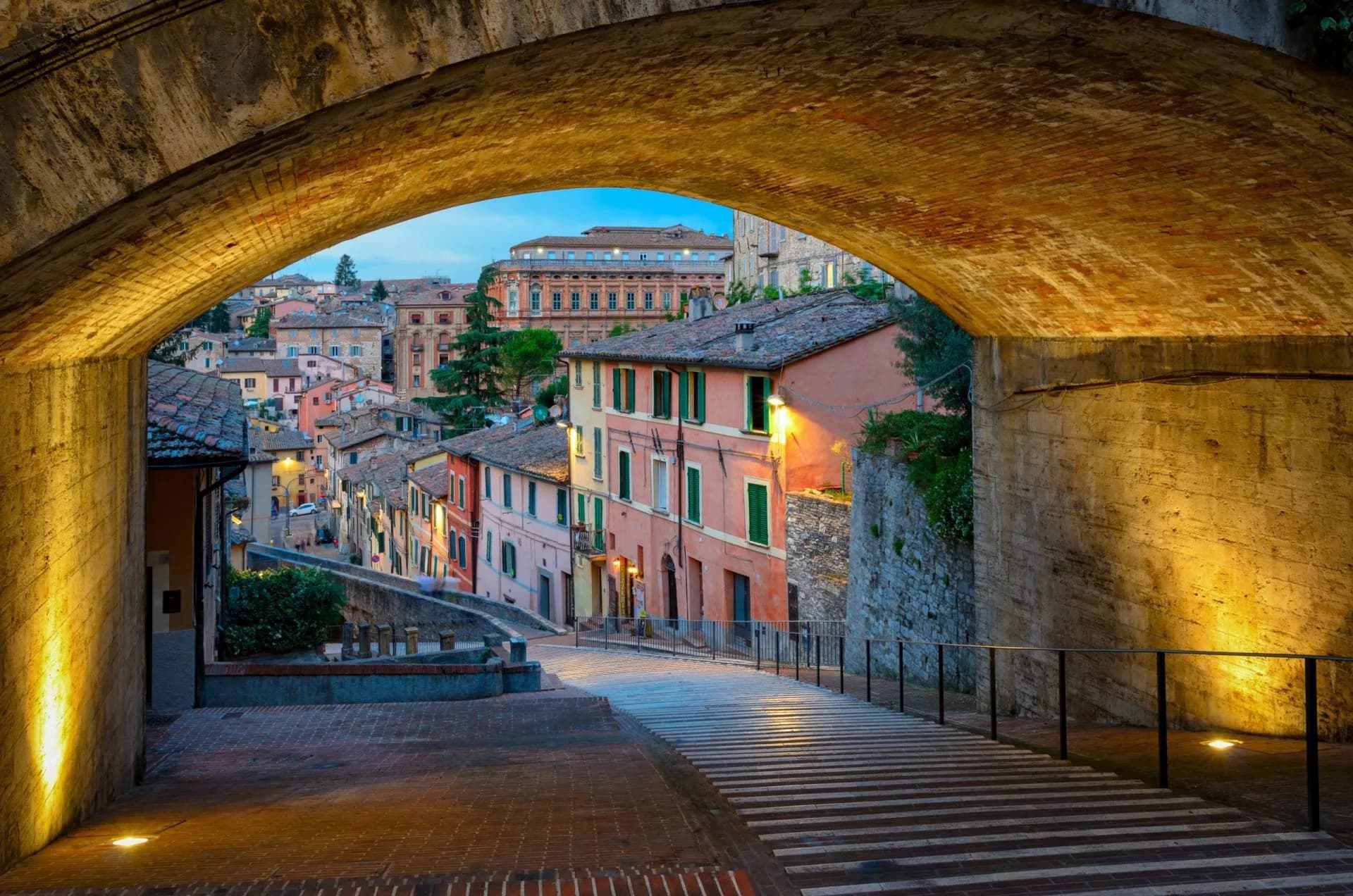 Historic Umbria town street viewed through a brick archway at dusk with illuminated buildings.