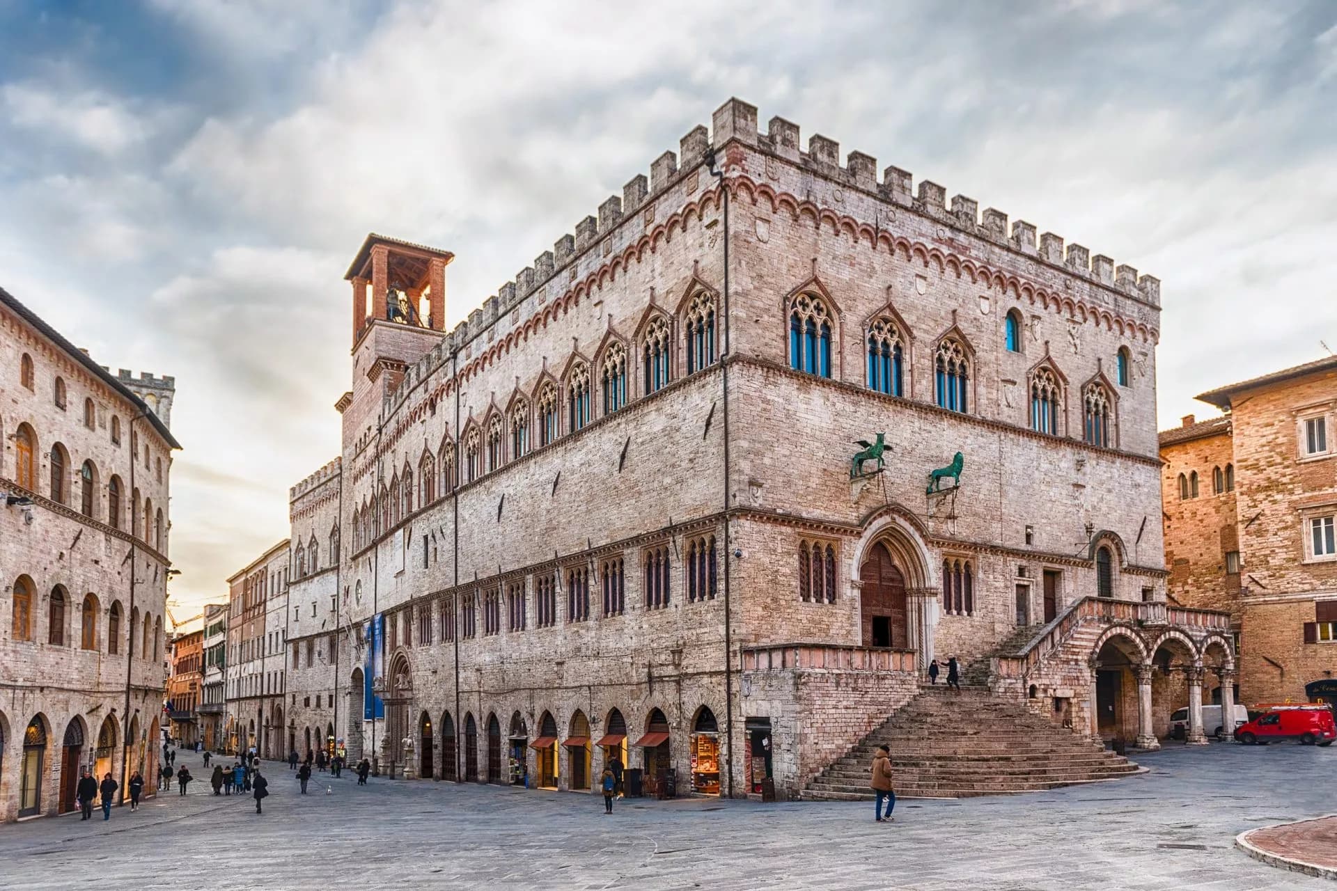 Palazzo dei Priori in a historic Italian square with people walking under a cloudy sky.