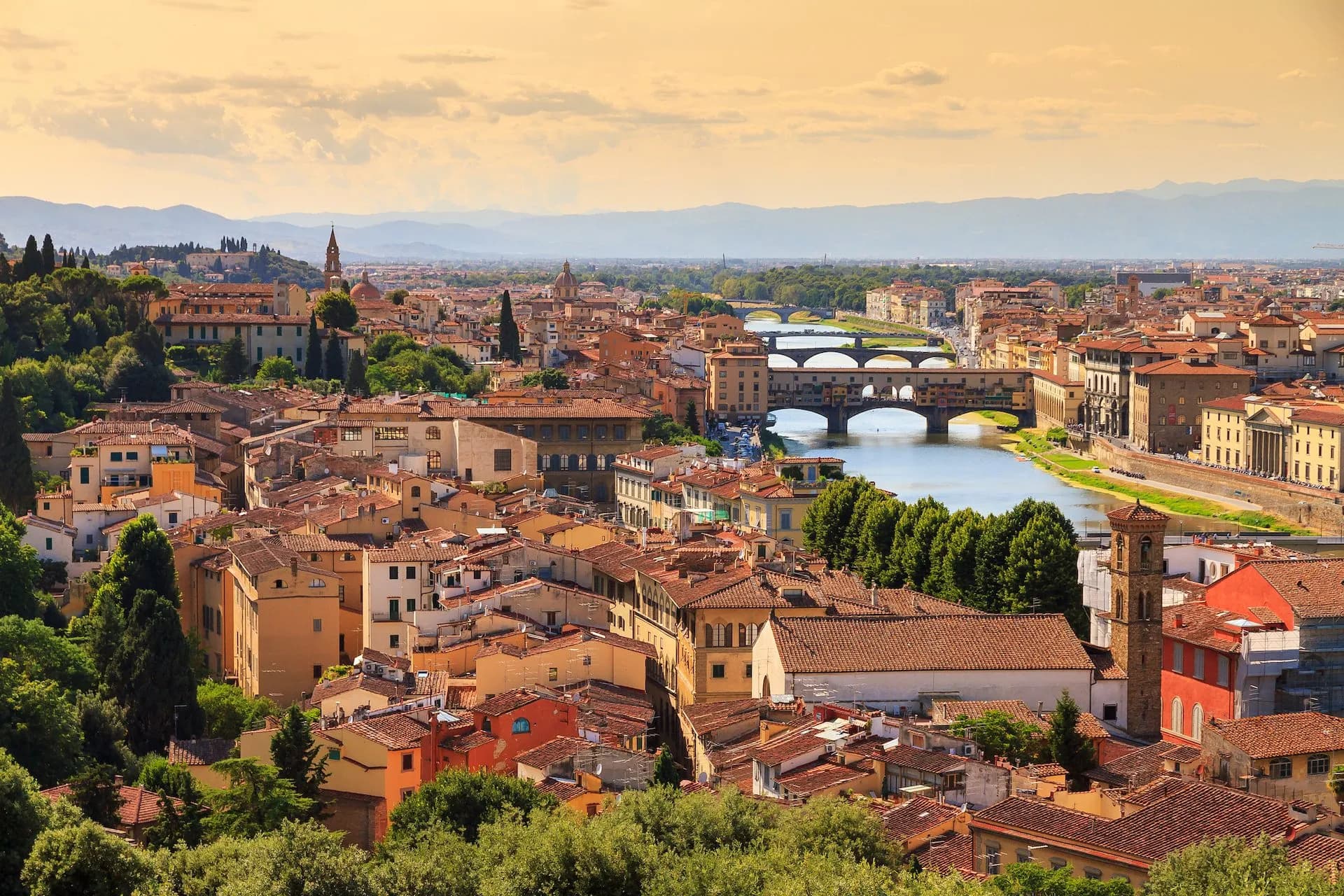 Florence cityscape with terracotta roofs, Arno River, and Ponte Vecchio bridge under a warm sky.