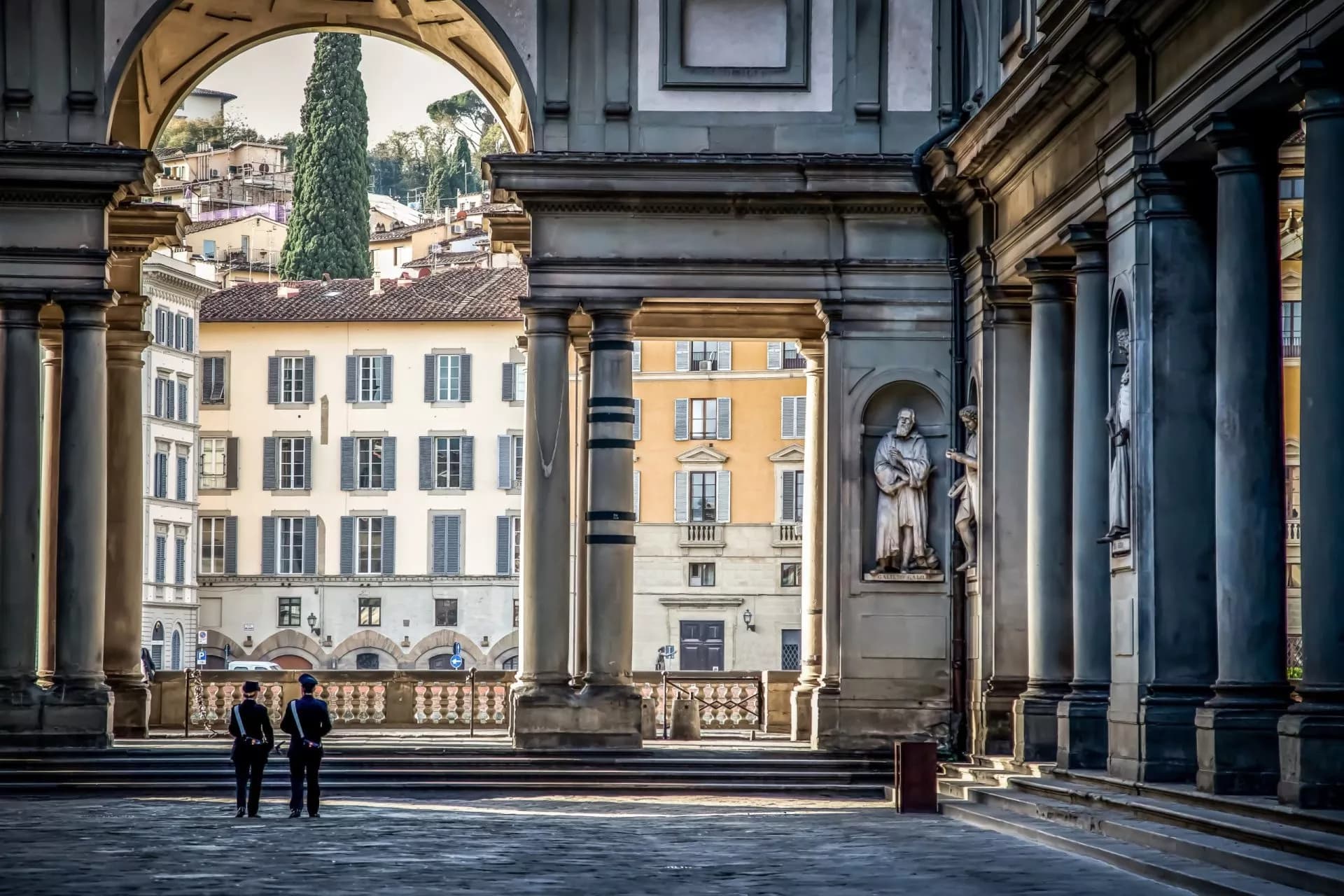 Uffizi Gallery loggia with statues, two guards, and view of Florence buildings.