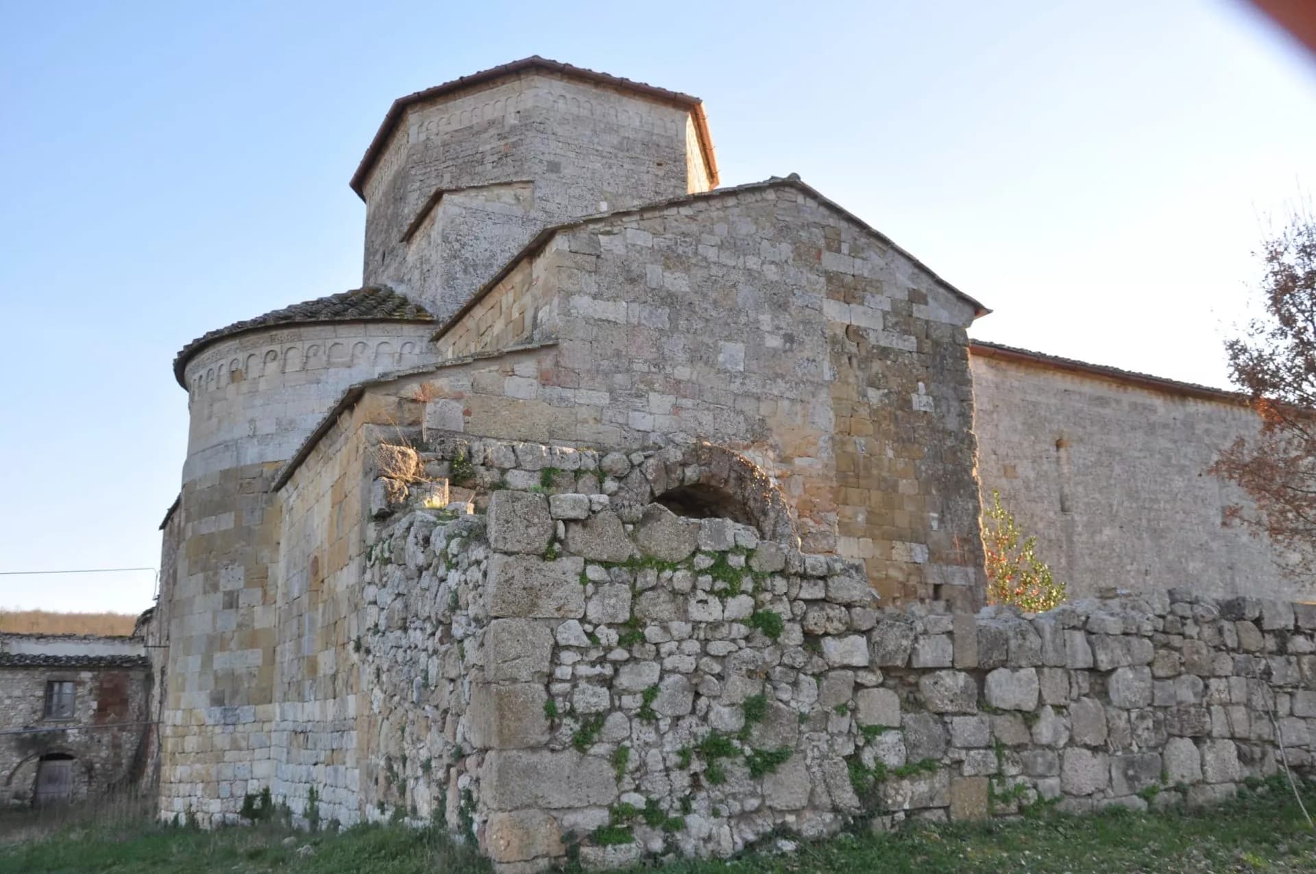 Abbey of Santa Maria a Conèo made of rough stone with a low stone wall in the foreground under a clear sky.