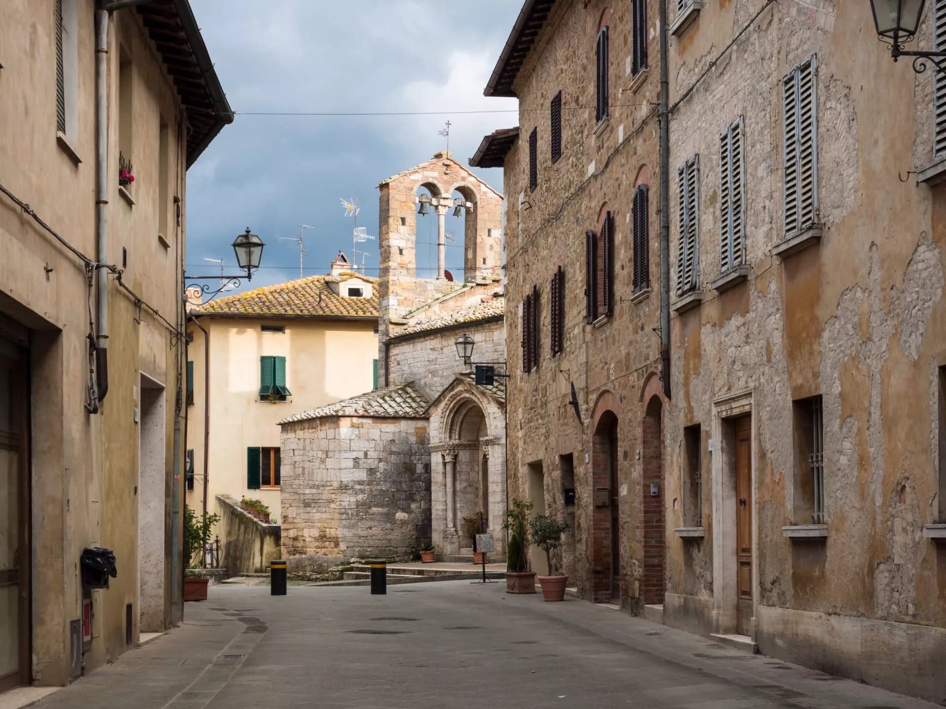 Narrow street in San Quirico d'Orcia leading to a stone church with a bell gable.