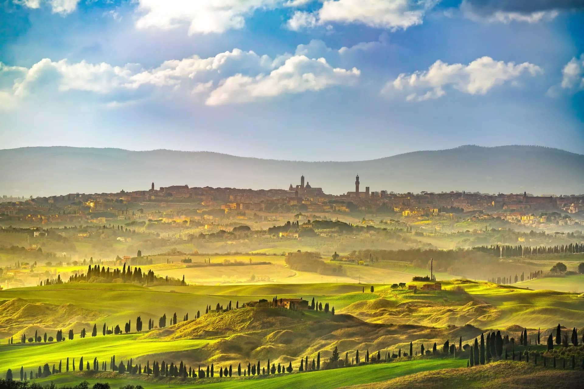 Siena skyline above rolling green Tuscan hills with cypress trees under a cloudy blue sky.