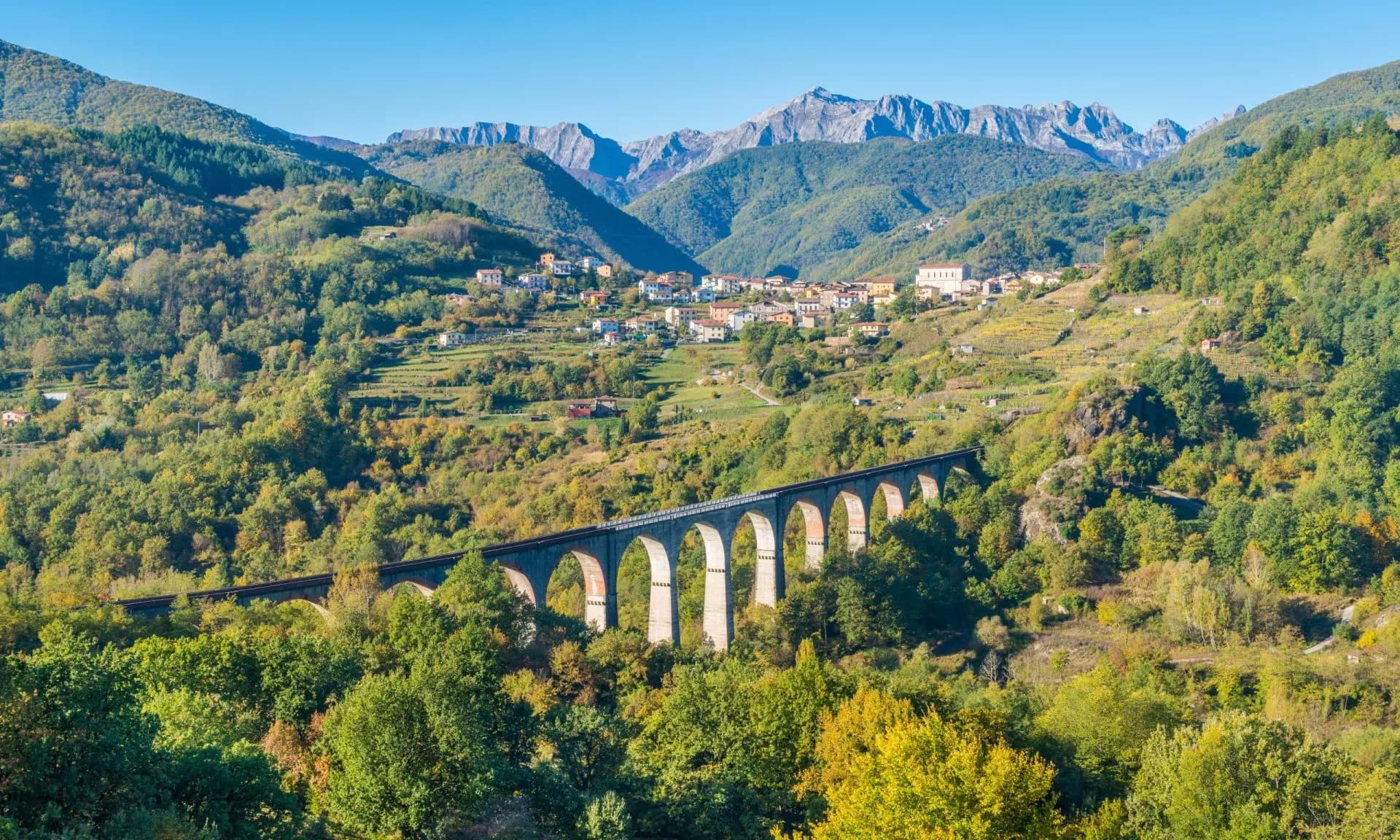 Viaduct crossing a lush valley with an alpine village and rugged mountains in the background.