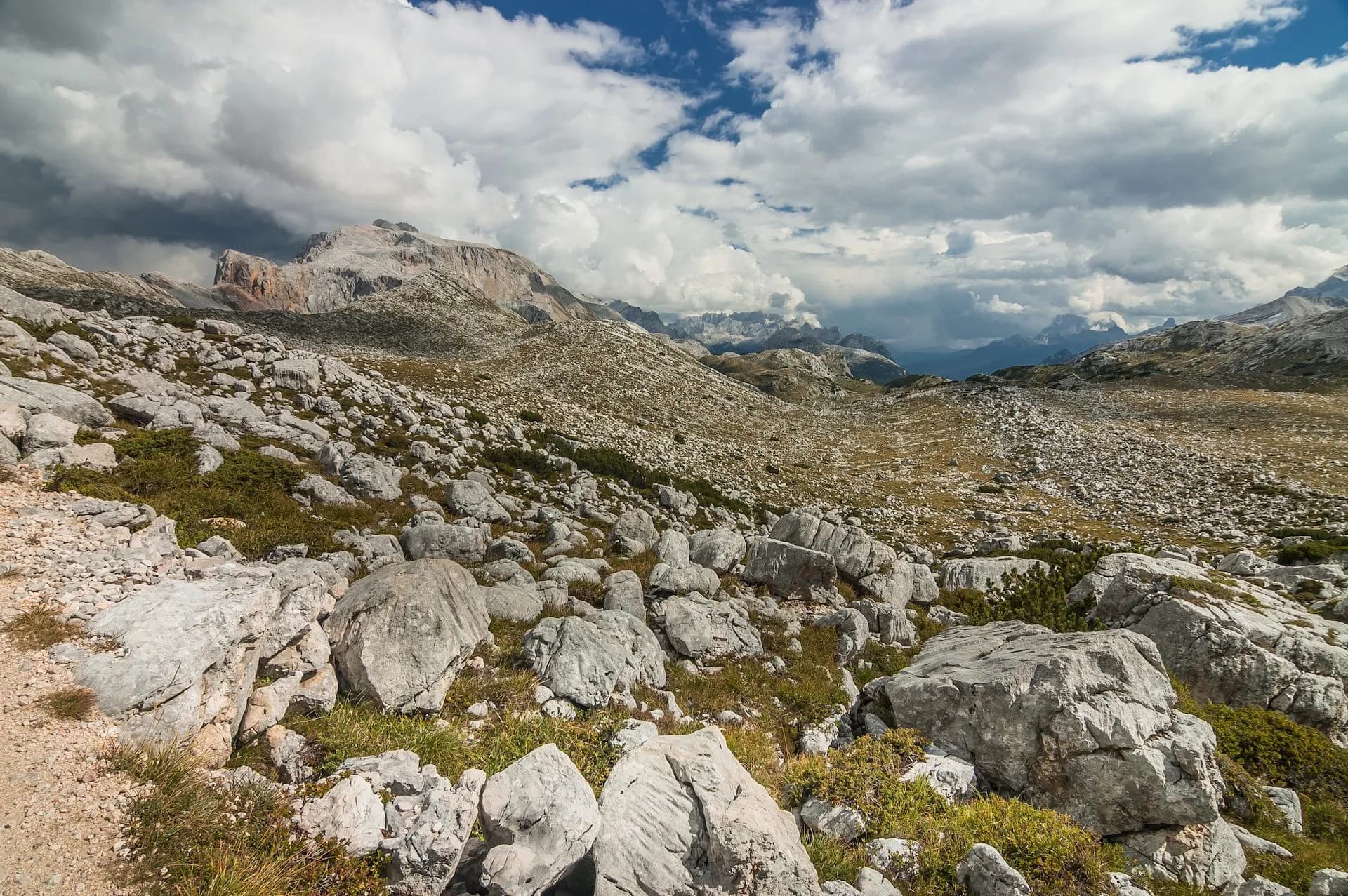 Rocky alpine terrain with sparse grass leading to distant mountain peaks under a cloudy sky near Rifugio Biella.