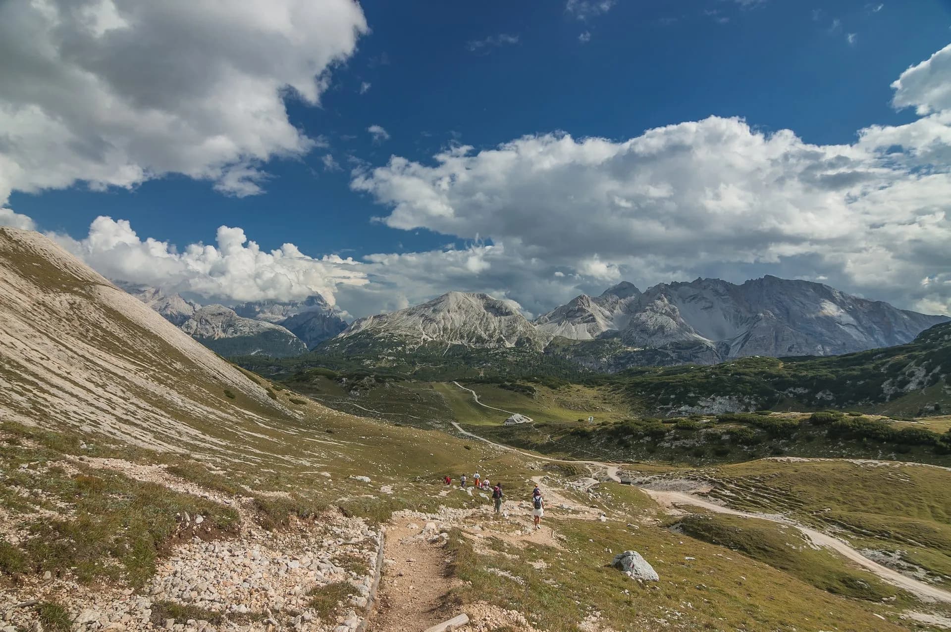 Hikers on rocky trail with dramatic mountains and cloudy blue sky, Alta Via 1.