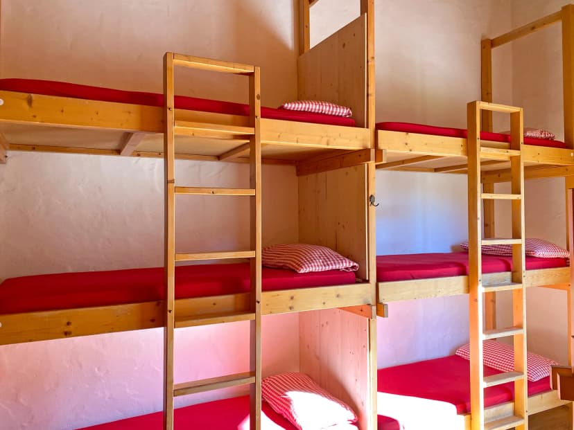 Wooden bunk beds with red mattresses and gingham pillows inside a simple room, likely a mountain hut.