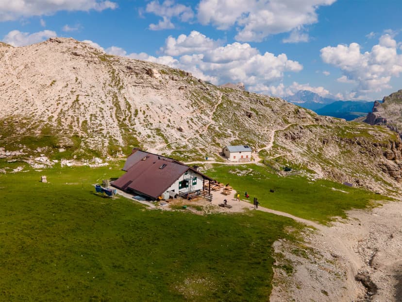 Mountain hut with brown roof on grassy slope near rocky peaks under blue sky, Puez-Hut.