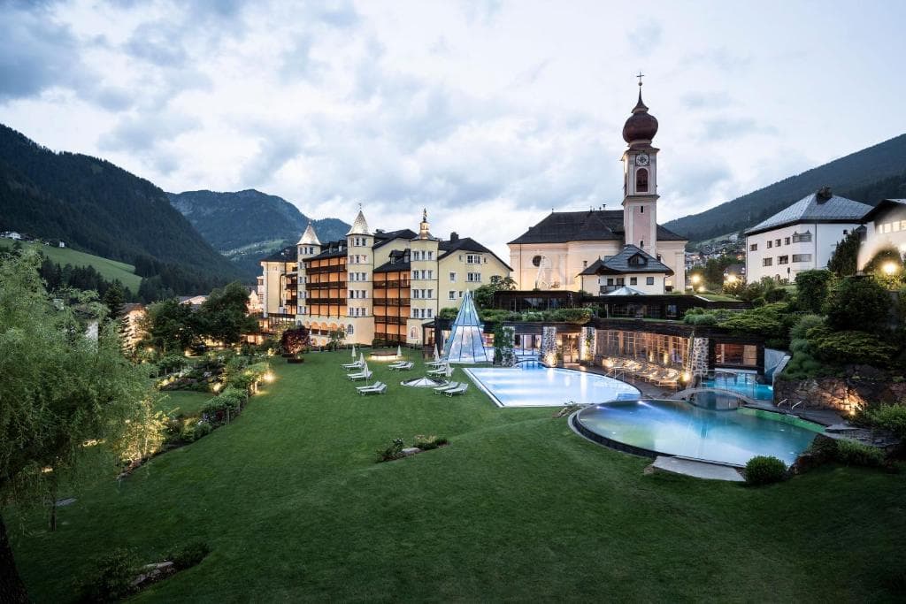 Resort hotel with outdoor swimming pools and church tower in an alpine mountain valley.