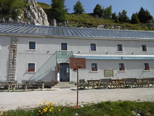 Mountain refuge building with outdoor seating and a sign pointing to a museum and shelter in an alpine setting.