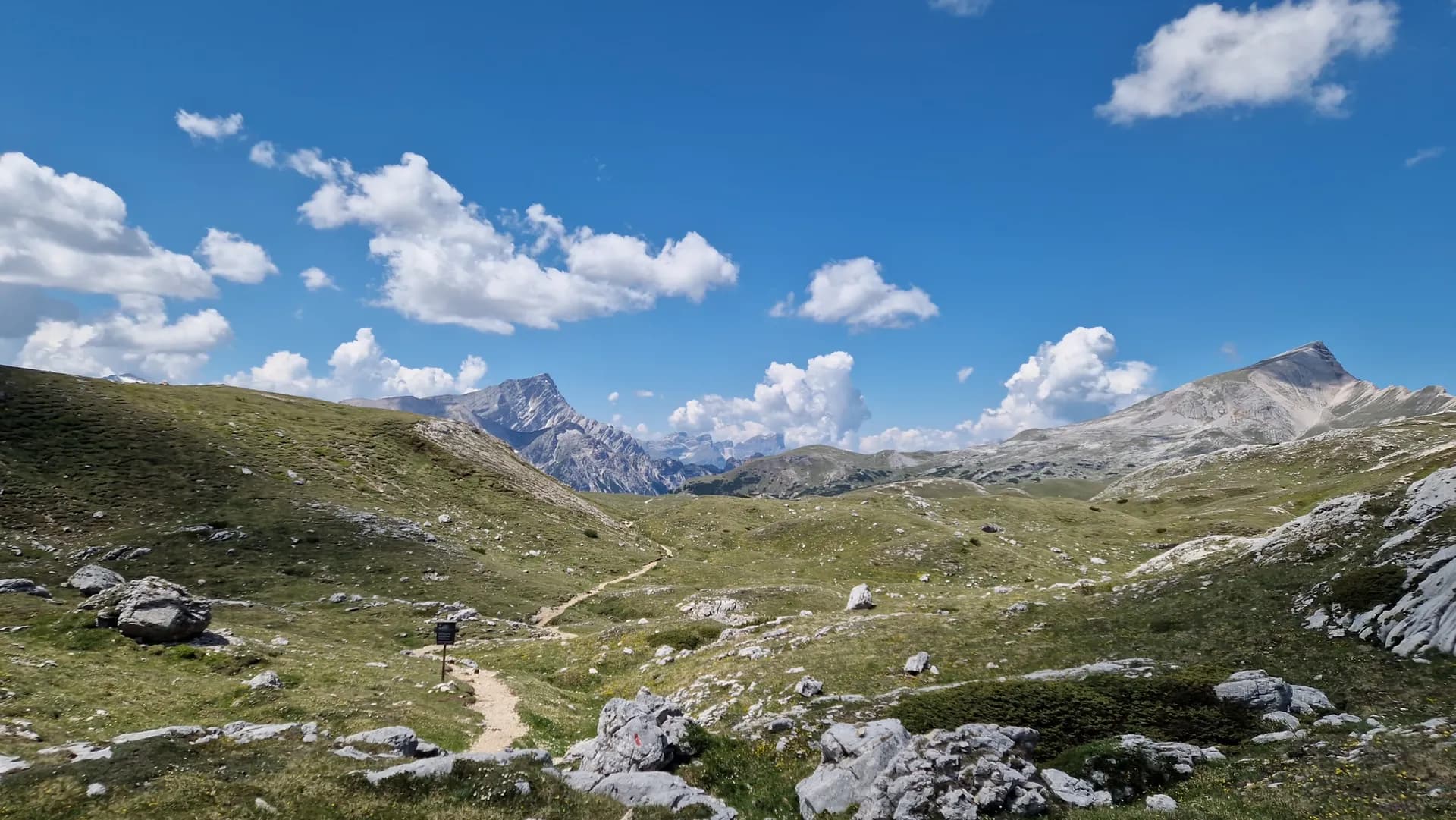 Hiking trail through a grassy alpine mountain valley under a blue sky with scattered clouds.