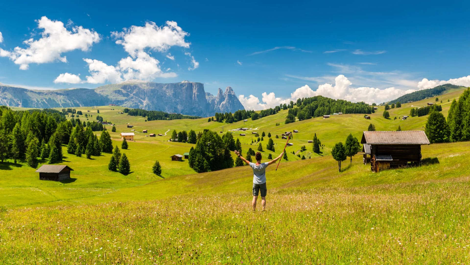Hiker with trekking poles enjoying sunny day on green Seiser Alm meadow with alpine huts and mountains.