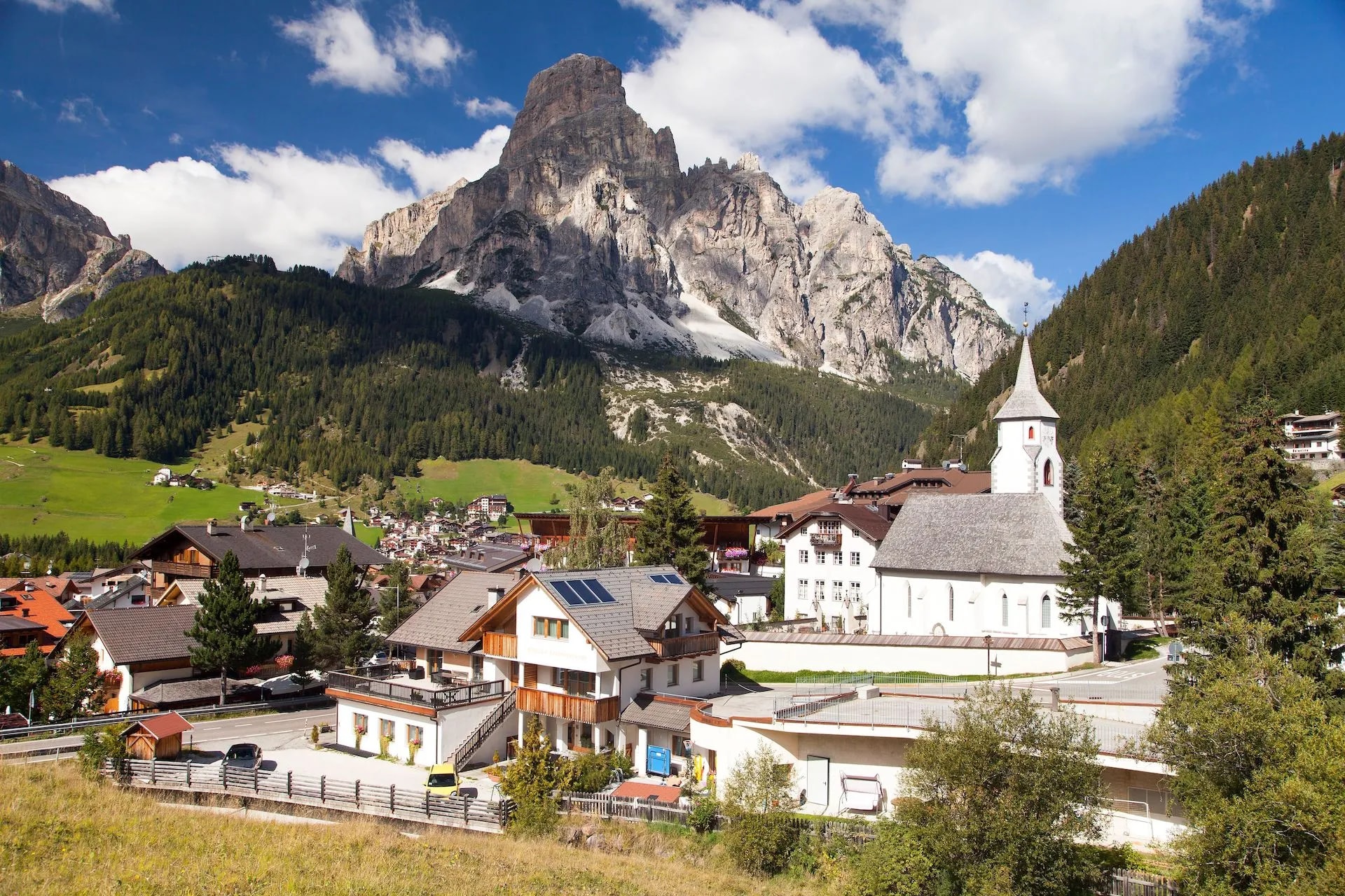 Alpine village of Corvara nestled below massive rocky peaks and green forested slopes.