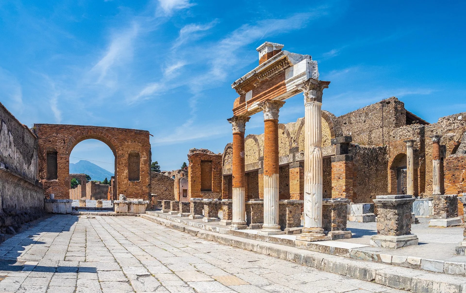 Ancient ruins of Pompeii with stone pathway, columns, and Mount Vesuvius visible through an arch.