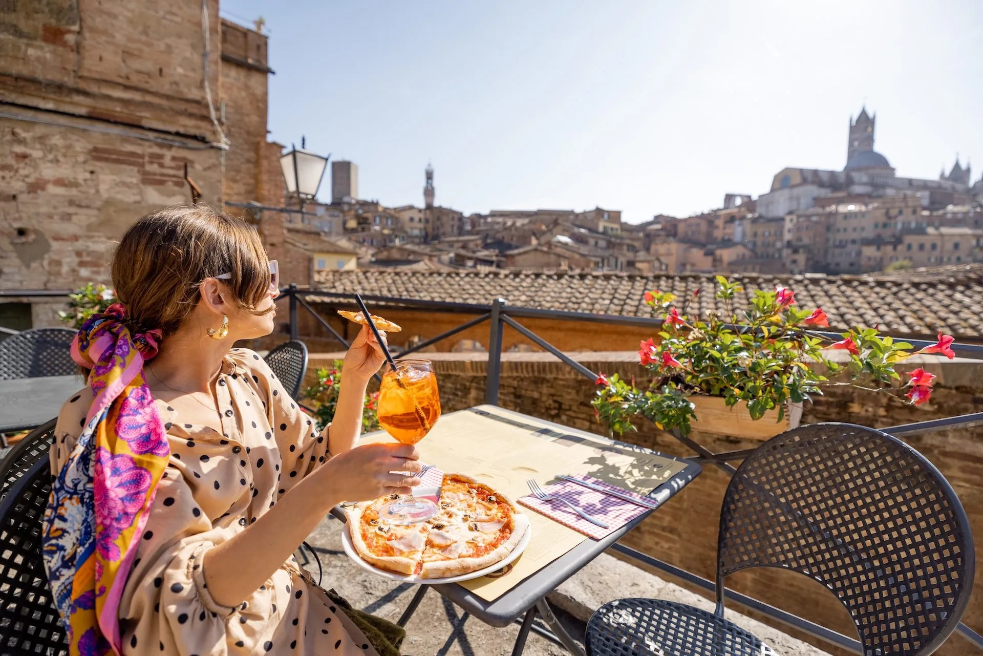Woman dining on pizza and Aperol Spritz on terrace overlooking Siena cityscape and Duomo.