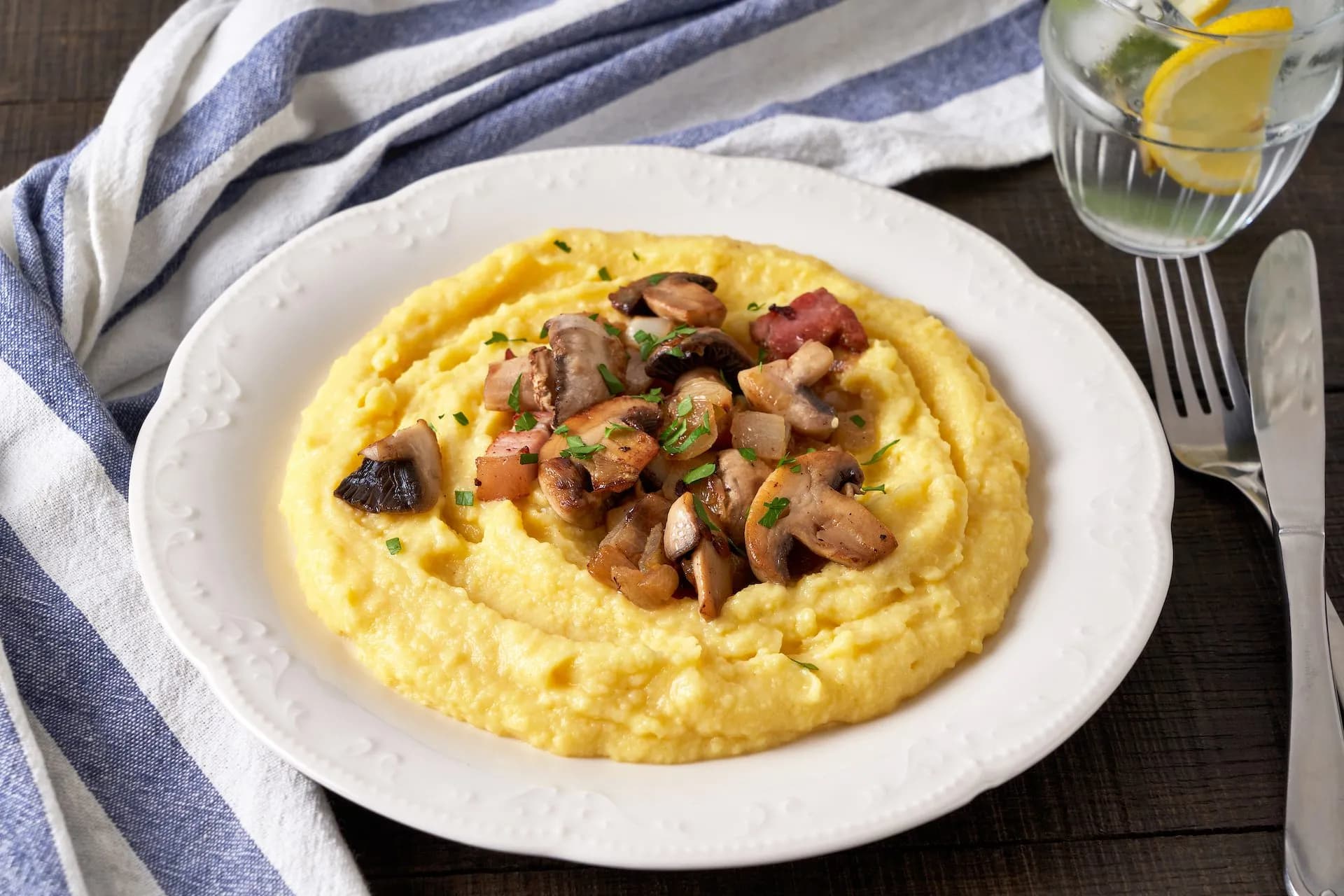 Creamy polenta topped with sautéed mushrooms and bacon on a white plate with silverware.