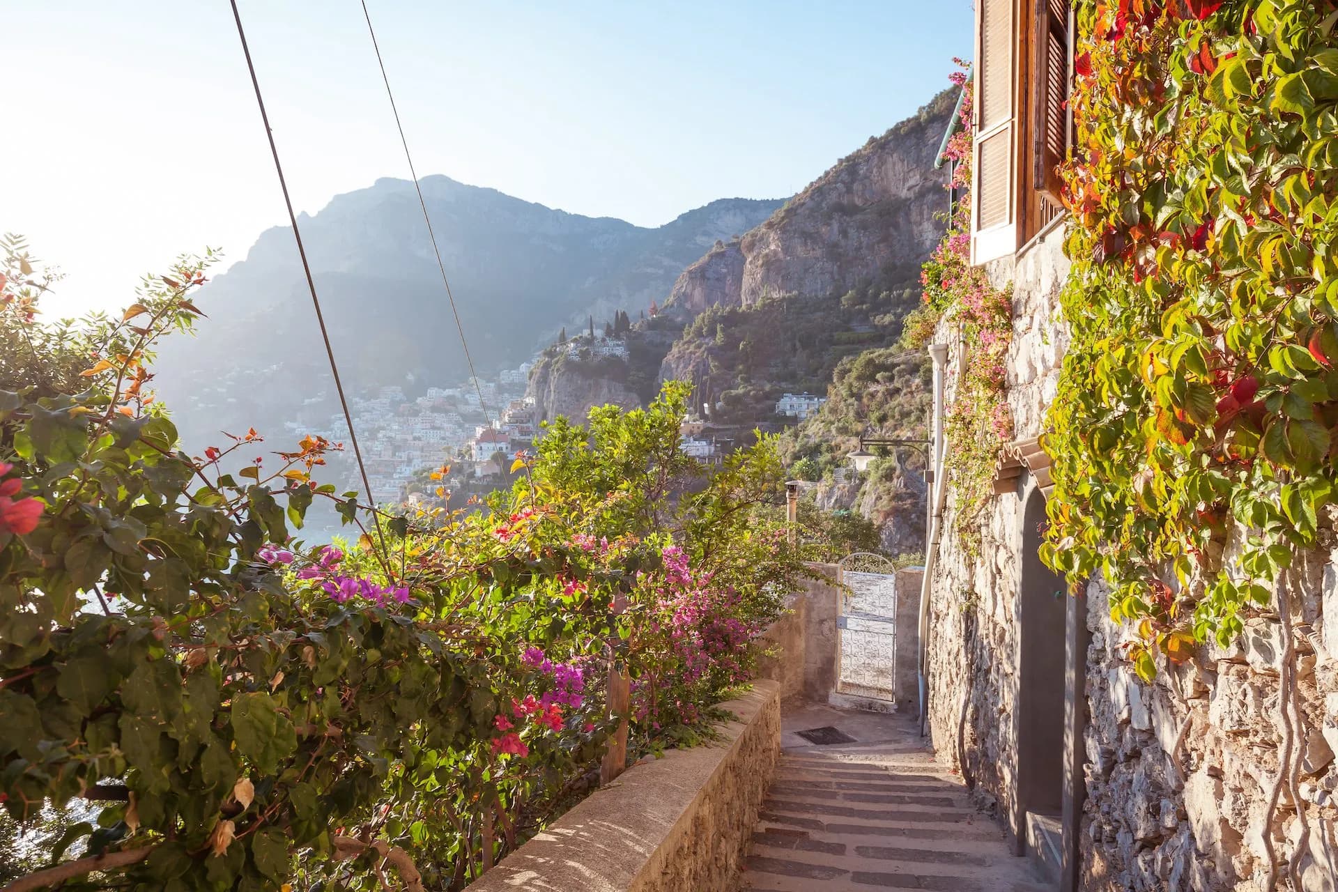Stone staircase path with flowering vines overlooking a coastal town nestled in steep mountains.