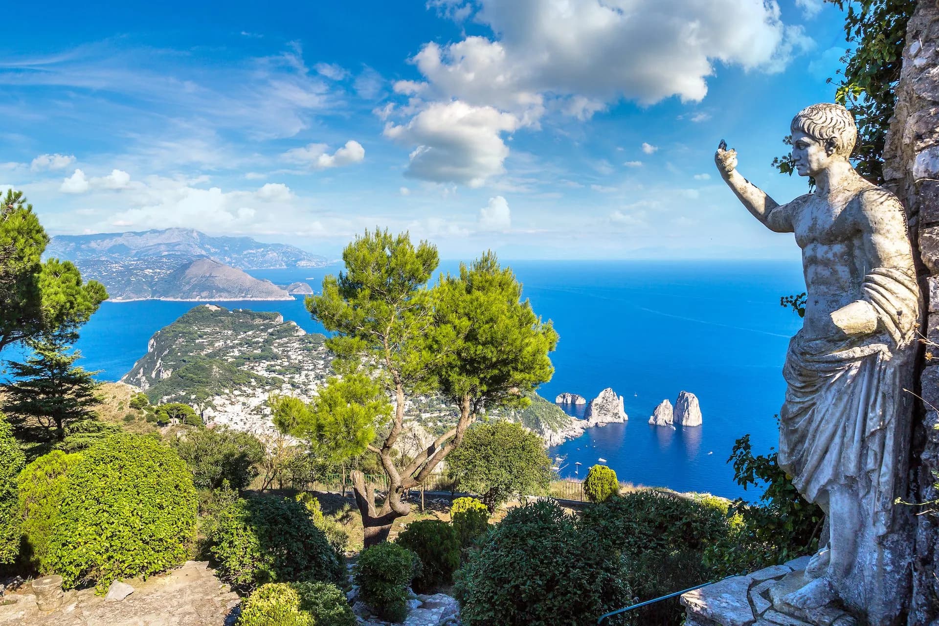 Capri view with statue, lush greenery, Faraglioni rocks, and bright blue Mediterranean Sea.