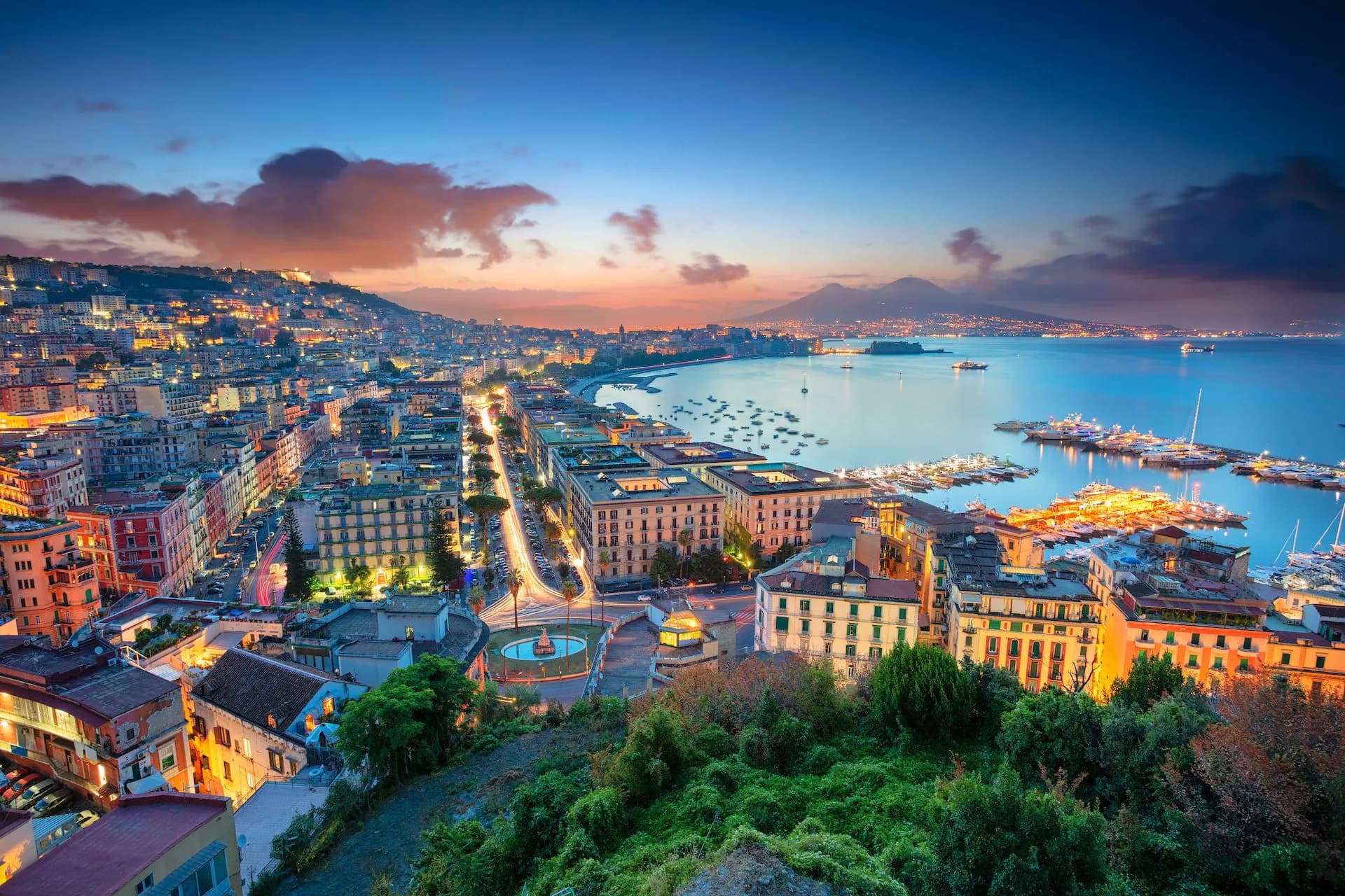 Cityscape of Naples at dusk with illuminated buildings, harbor, and Mount Vesuvius in the background.