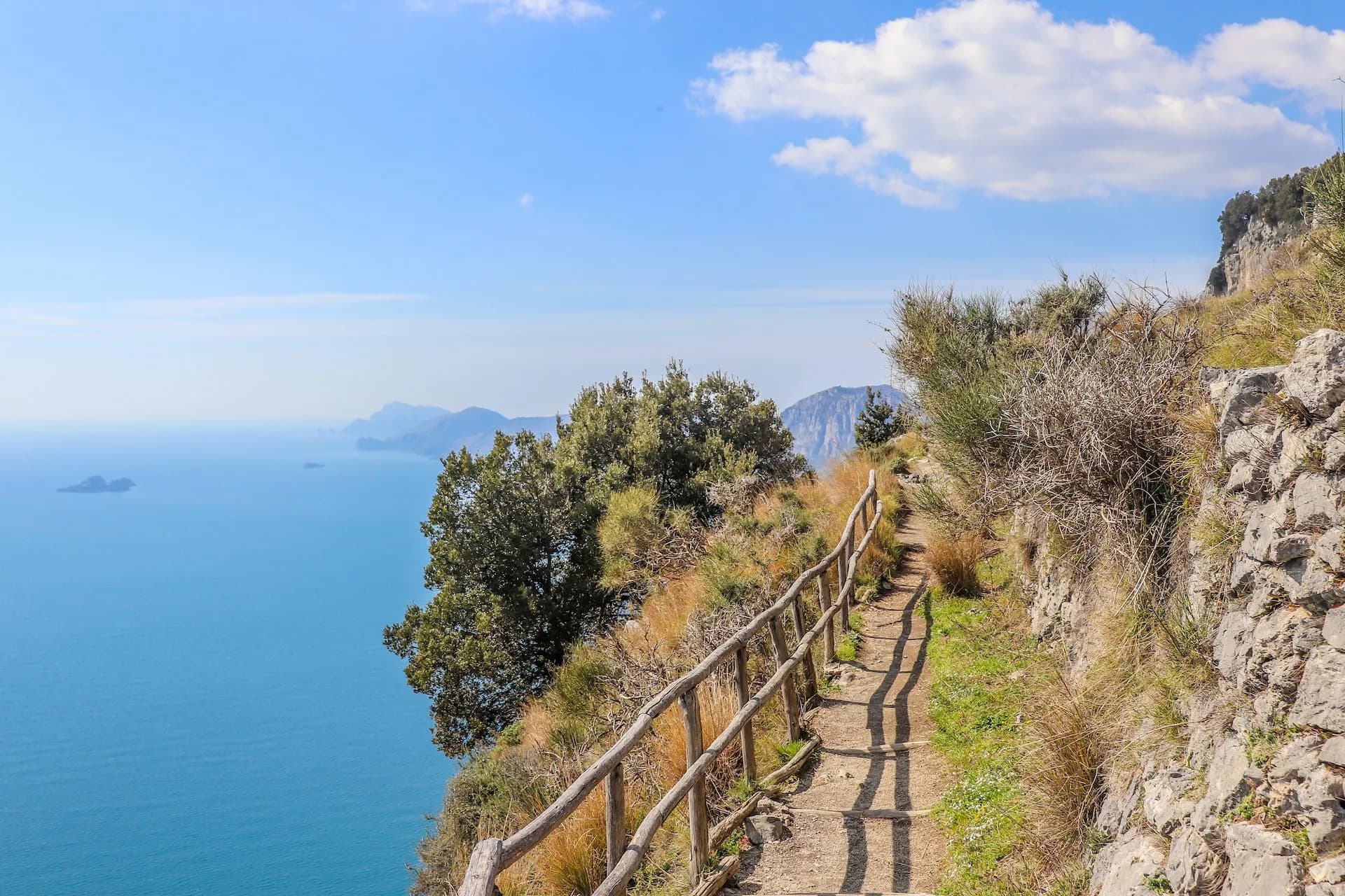 Hiking path along a cliff edge with wooden railing overlooking bright blue sea and distant mountains