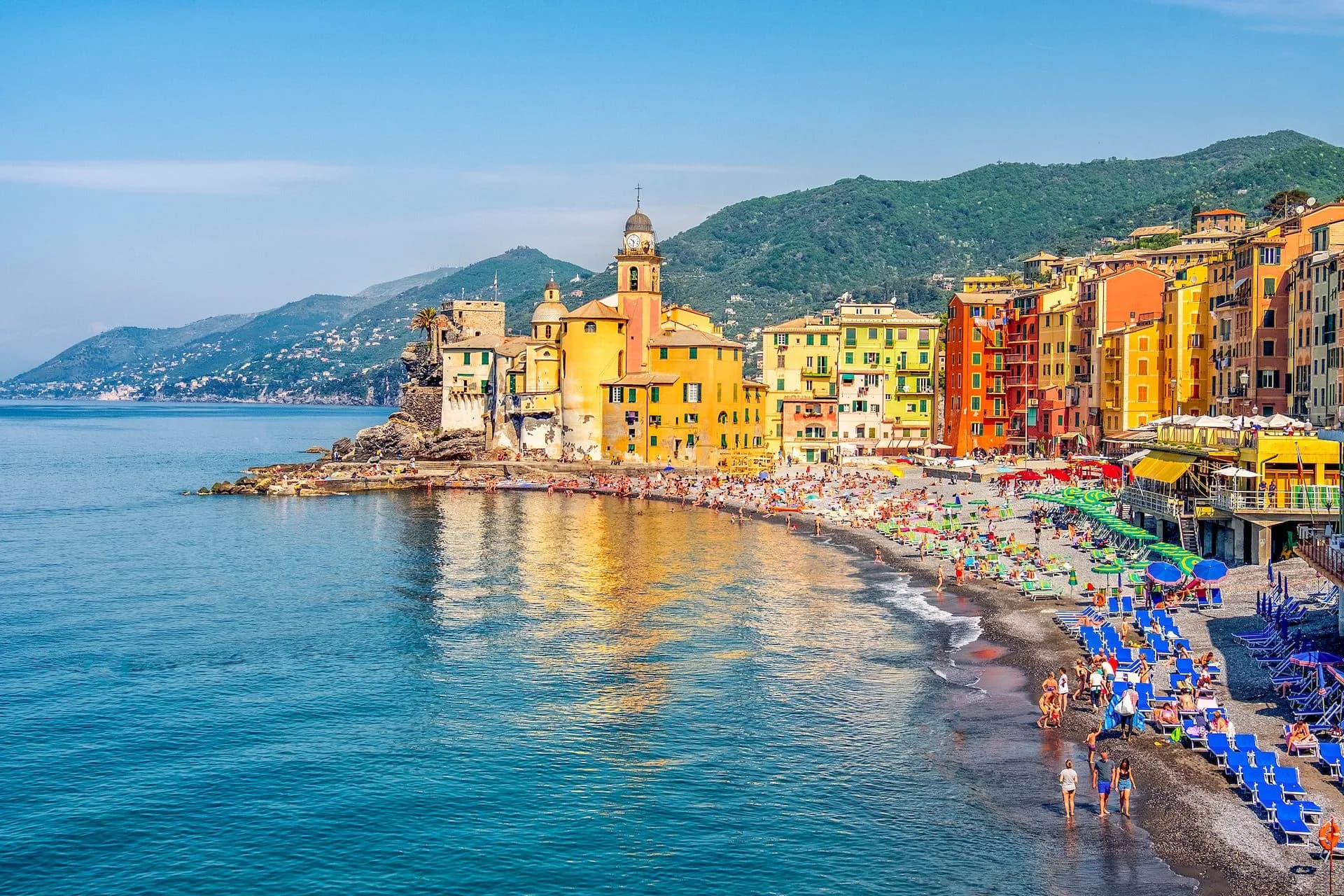 Colorful buildings line the beach in Camogli with sunbathers and green hills in the background.