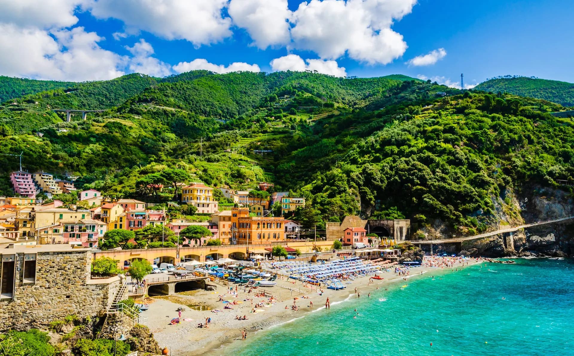 Beachgoers on a pebble beach next to turquoise water below colorful buildings and green hills in Monterosso.