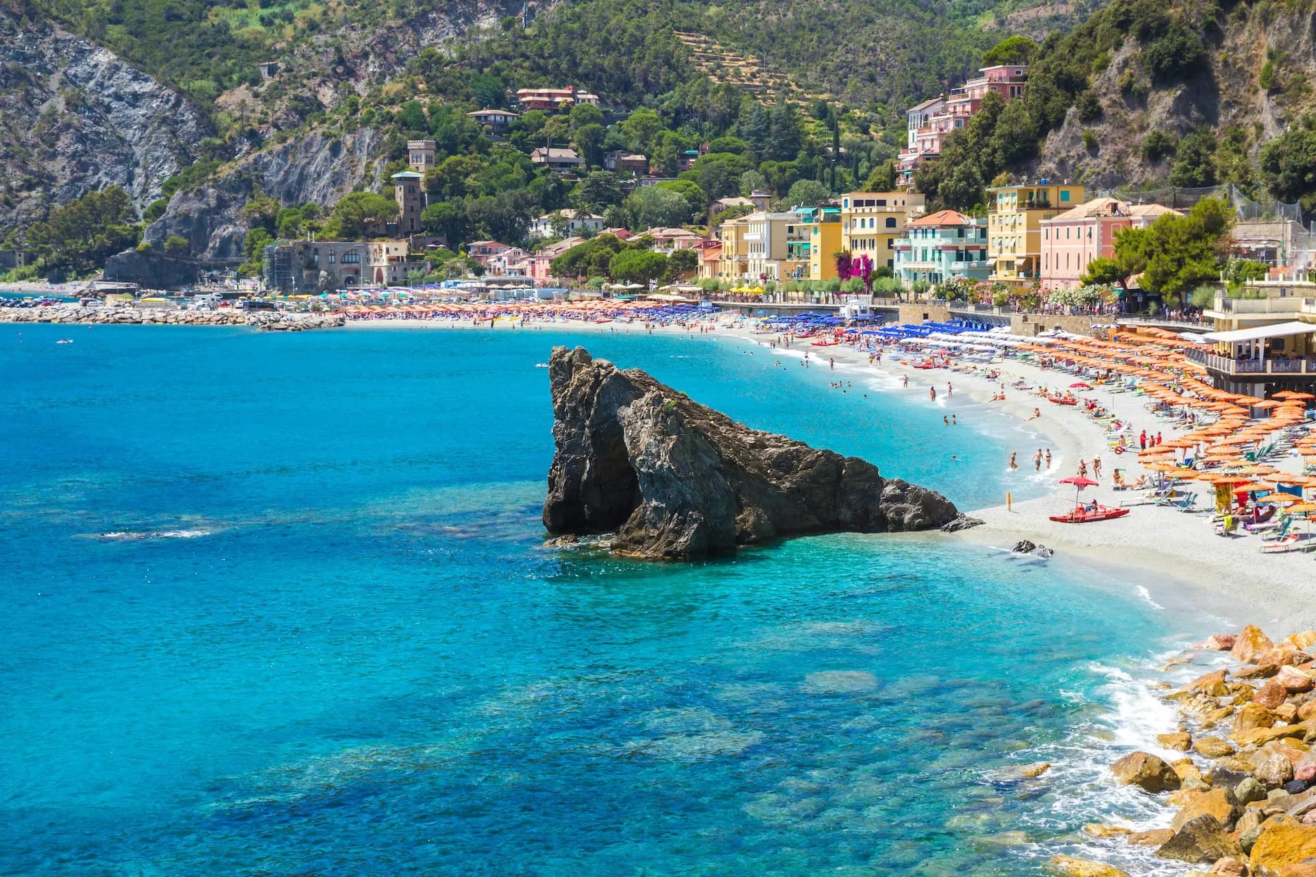 Beach crowded with orange umbrellas, turquoise water, and colorful buildings on a steep hillside in Monterosso.