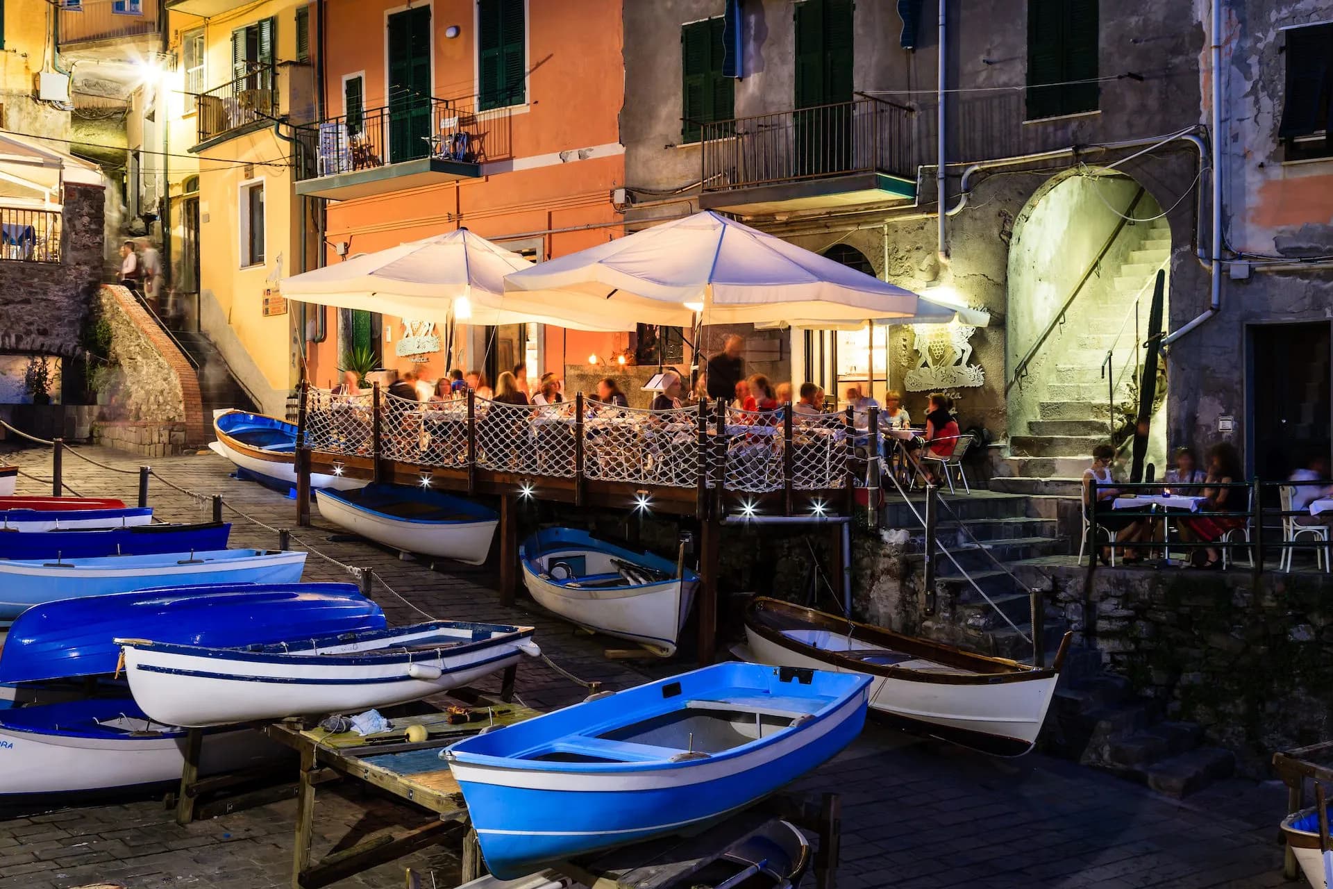 Outdoor dining terrace next to moored blue and white boats in Riomaggiore at night.