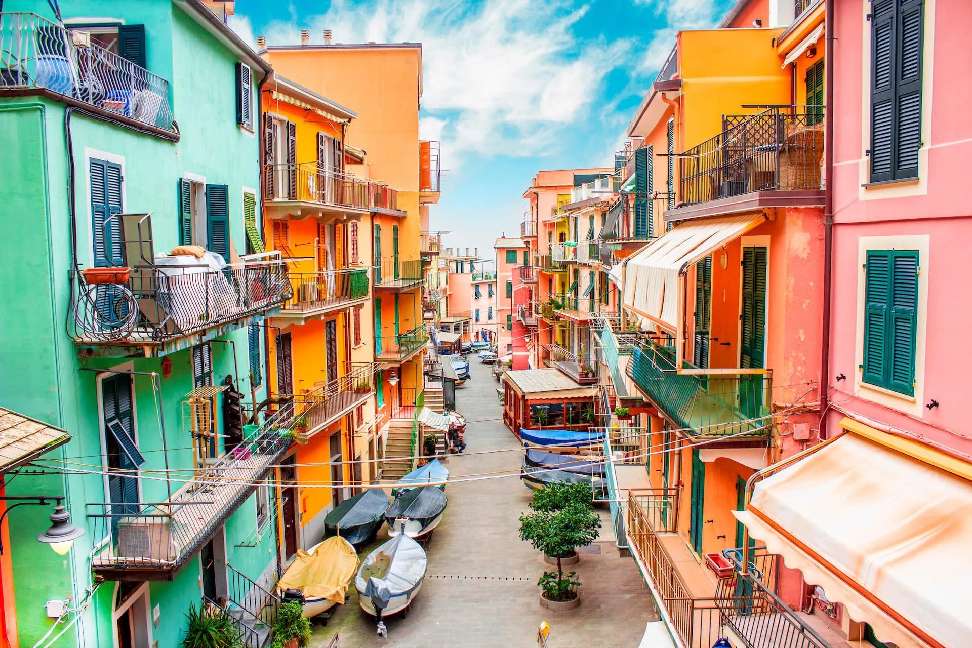 Colorful buildings line a narrow street with covered boats parked under a bright blue, cloudy sky in Manarola.