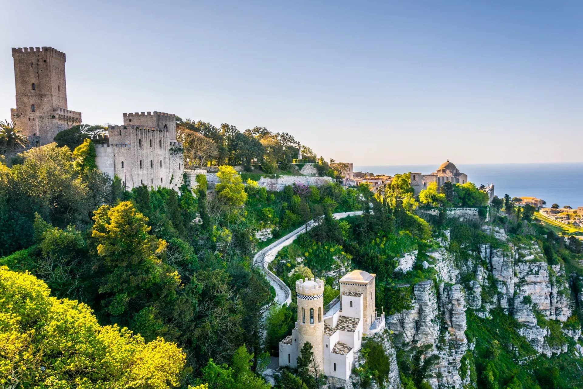 Historic stone castles and buildings perched on a lush green cliff overlooking the Mediterranean Sea in Erice, Sicily.