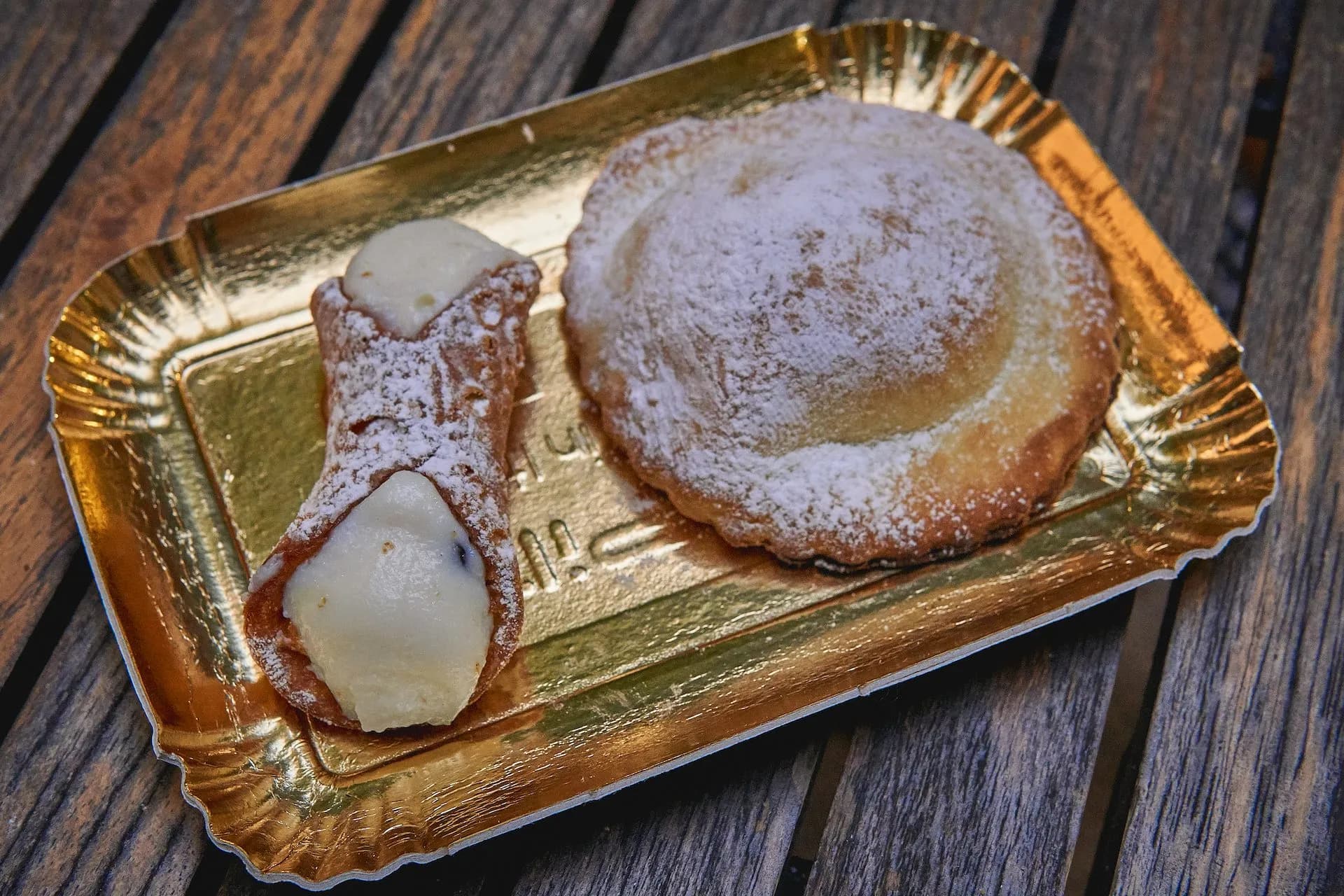 Cannoli and pastry dusted with powdered sugar on a gold tray on wood